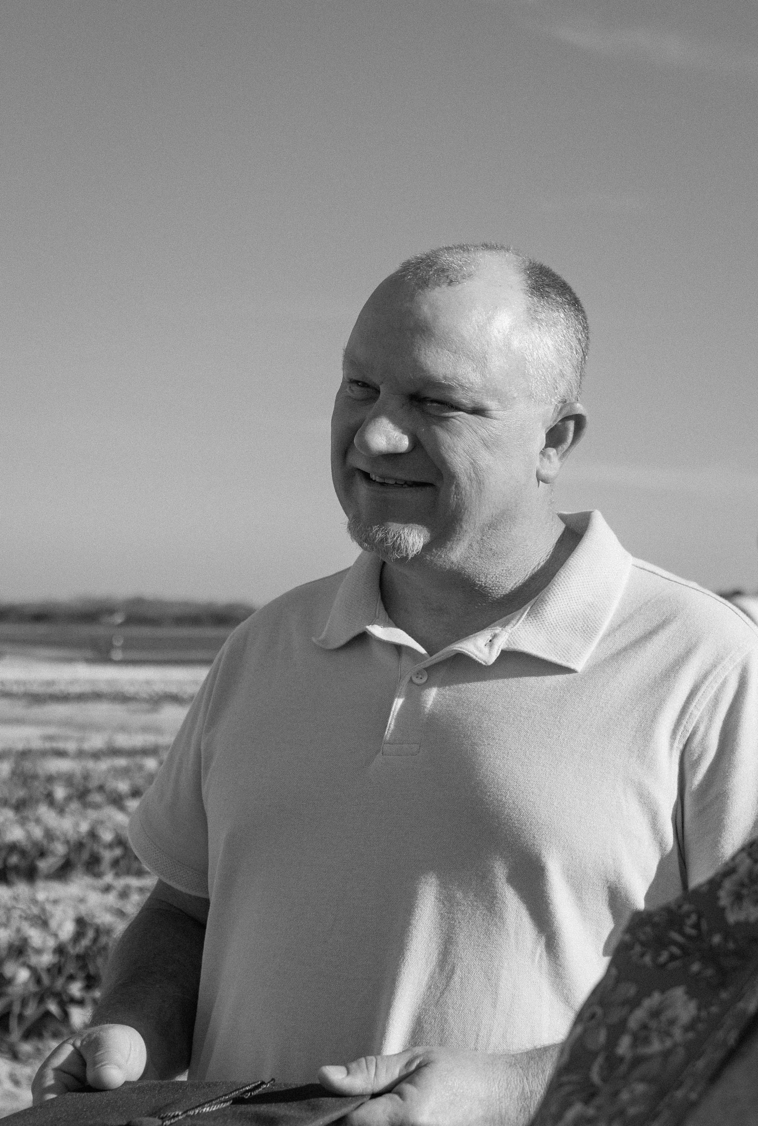 A man with a goatee and short hair, smiling, outdoors on a sunny day, wearing a light-colored polo shirt, with a landscape in the background.
