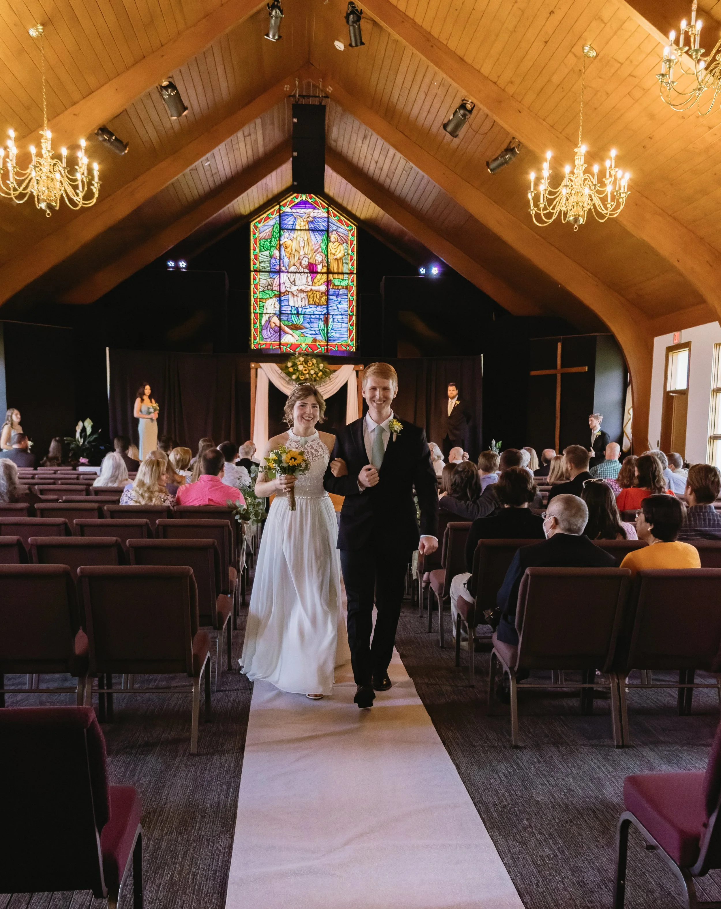 A newlywed couple walking down the aisle inside a church, with guests seated on either side, a stained glass window above the altar, and chandeliers hanging from the wooden ceiling.