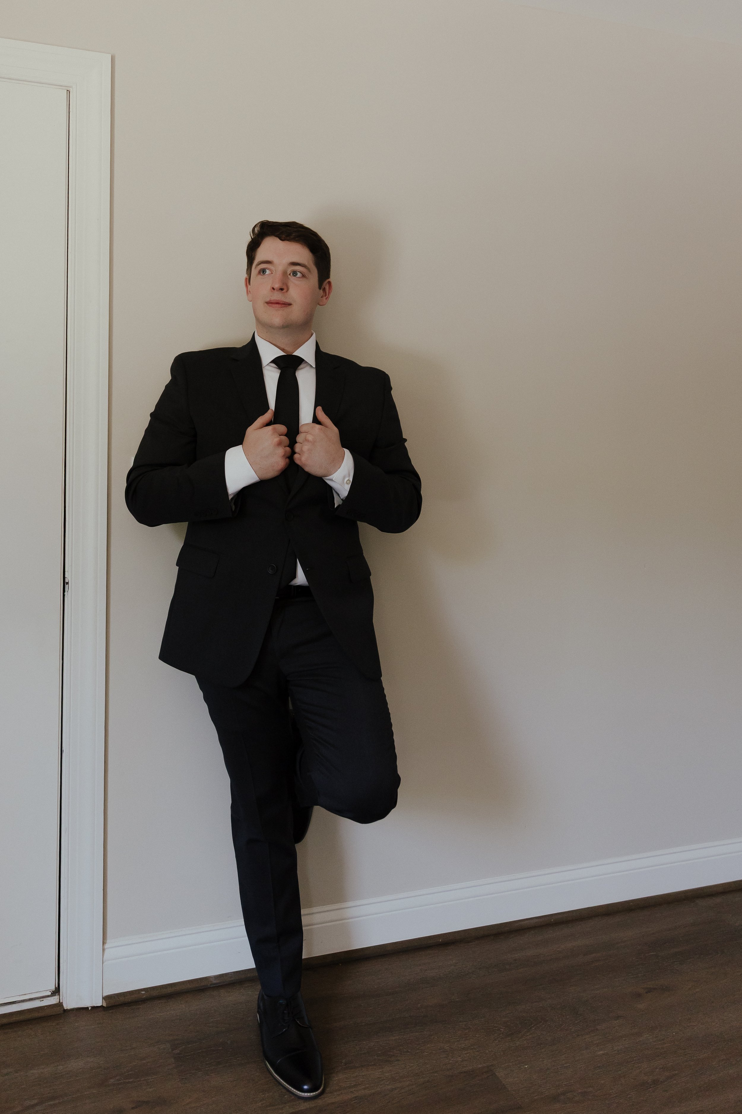 A photo of a groom leaning up against a wall, looking off into the distance. The groom is dressed in his suit and is holding the collar of his suit.