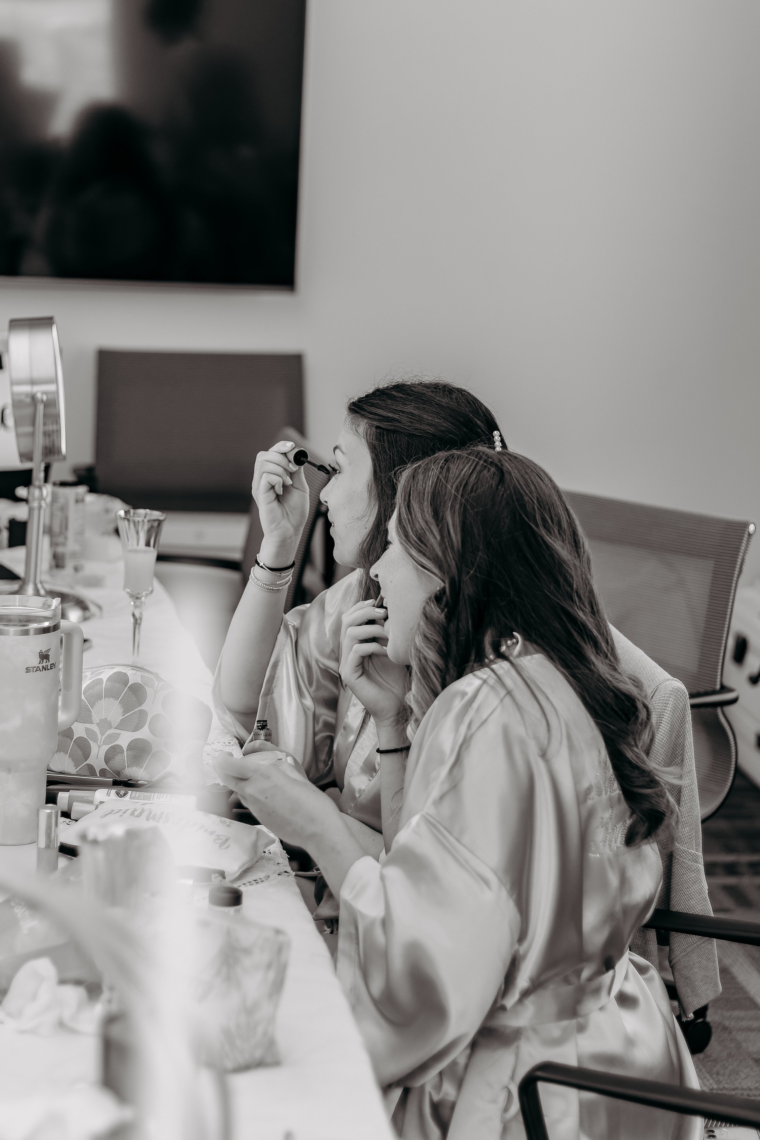 Two women in satin robes sit at a table, one applying makeup and the other looking at her phone, with makeup and beauty tools scattered on the table.