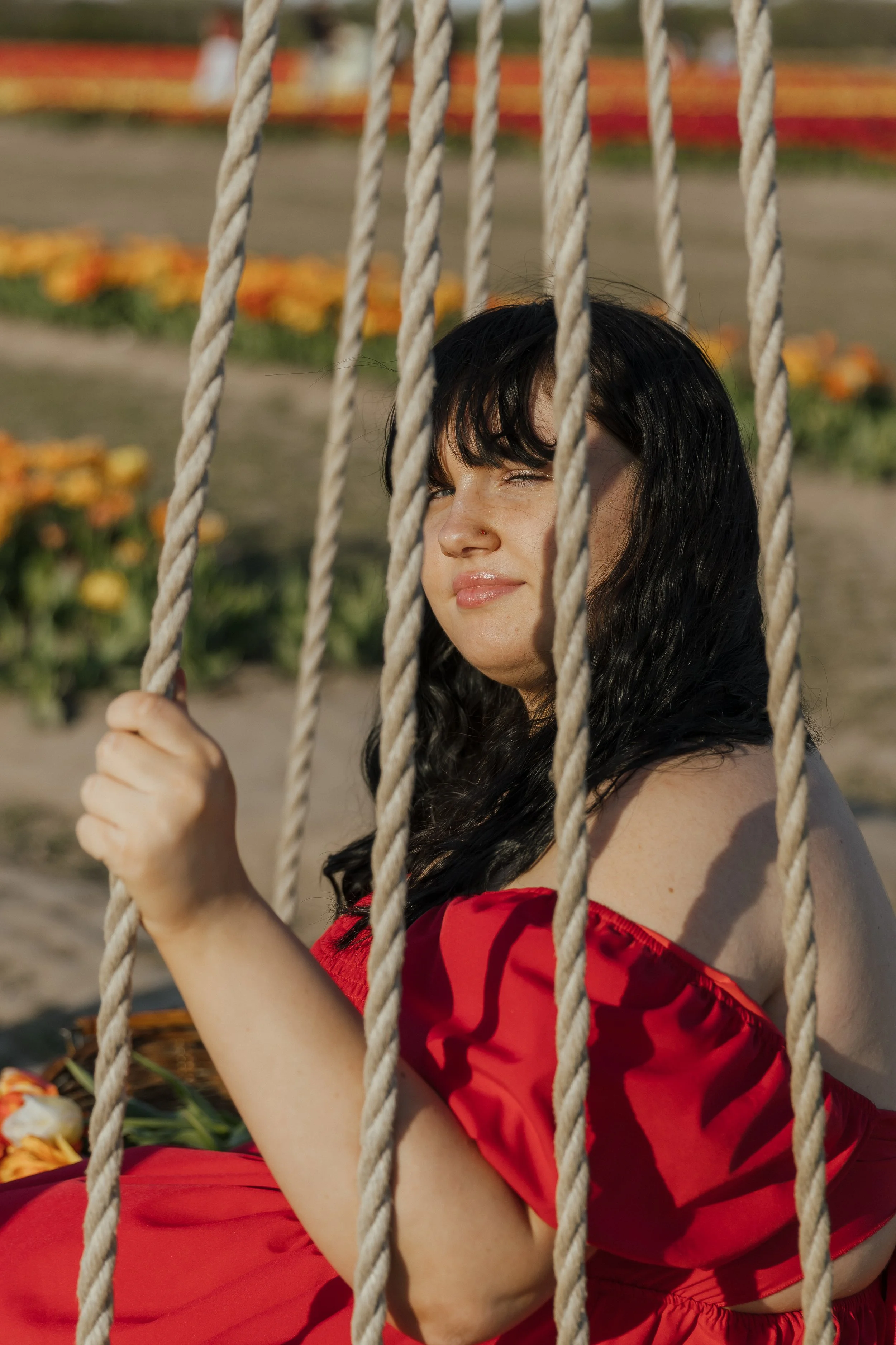 A woman with dark hair and light skin, sitting on a swing with thick rope, wearing a red off-shoulder dress, smiling slightly, with a floral garden in the background.