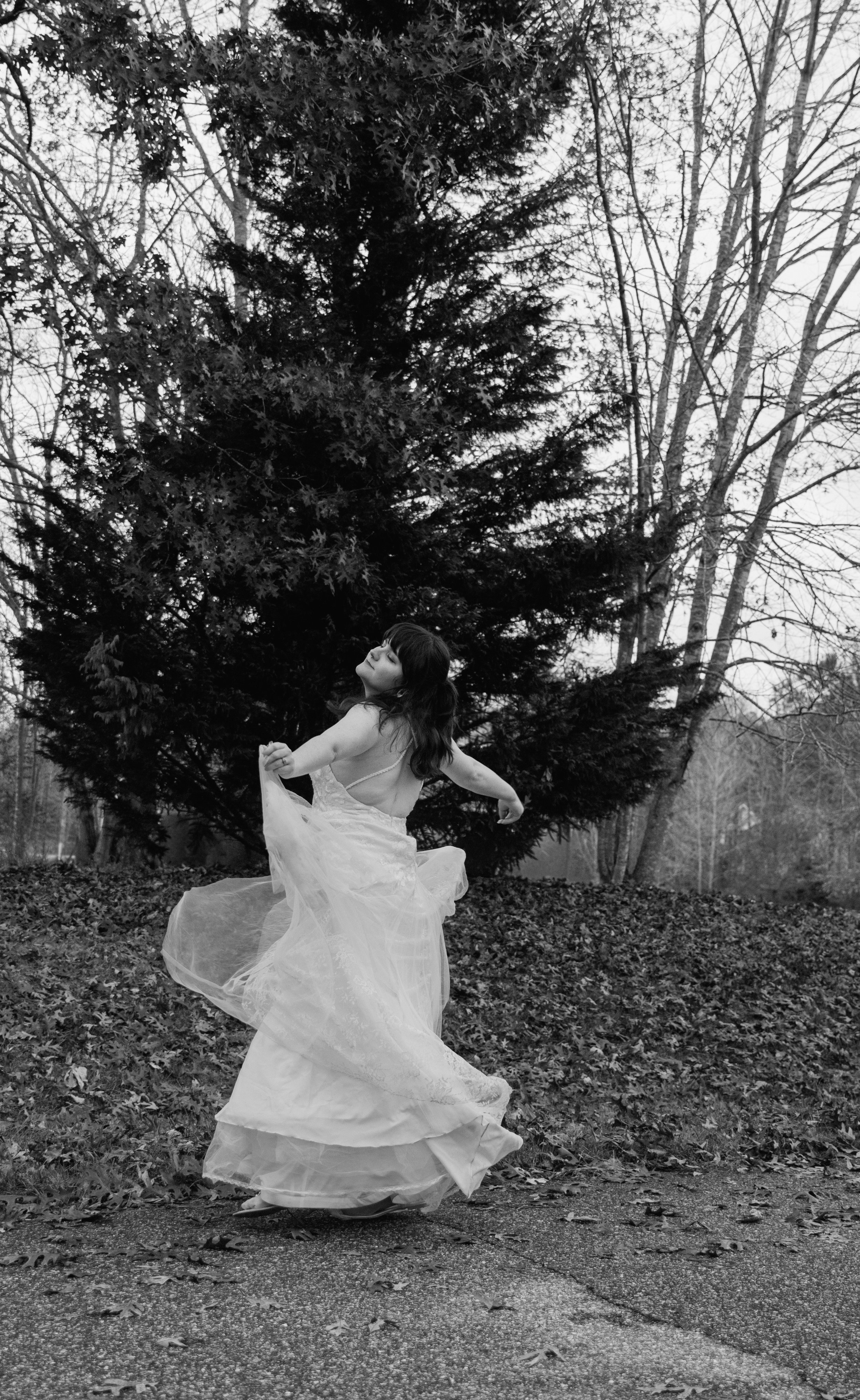 A woman in a flowing dress dancing outdoors on a paved surface, with trees and a large pine tree in the background, in black and white.