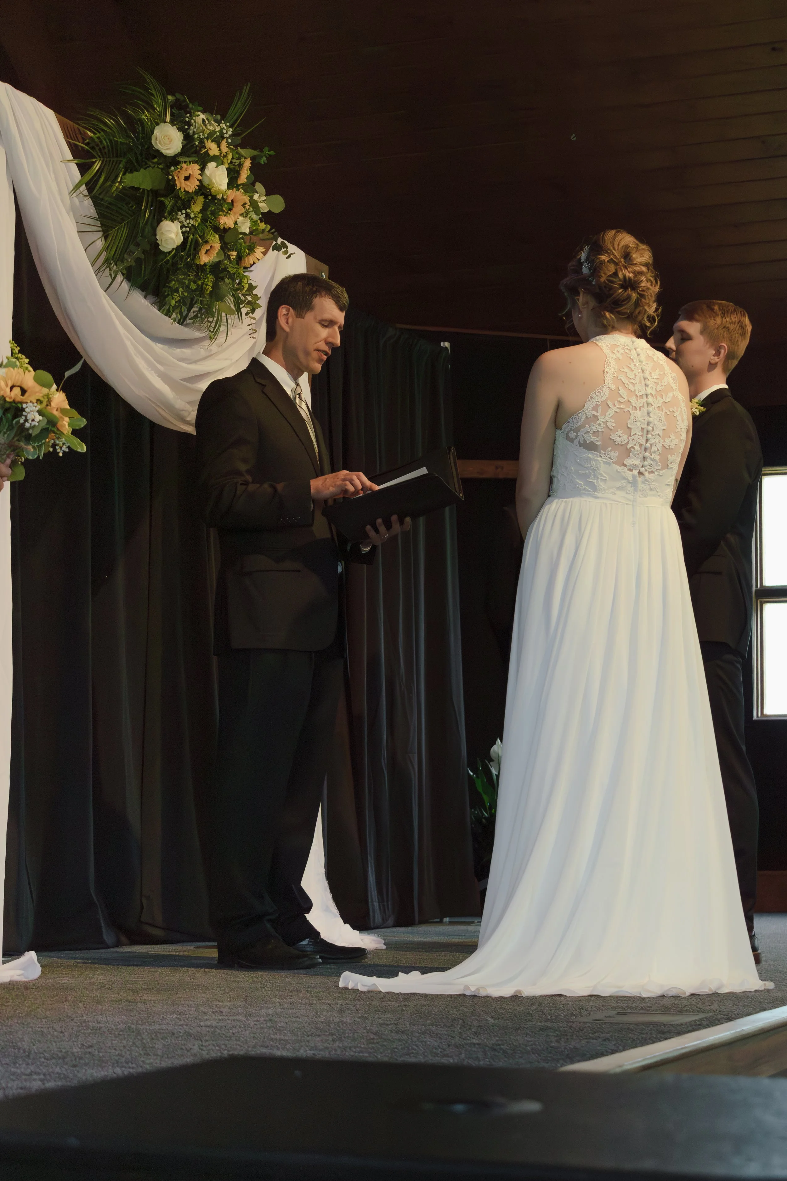 A couple getting married with an officiant during their wedding ceremony indoors, with floral decorations and black curtains in the background.