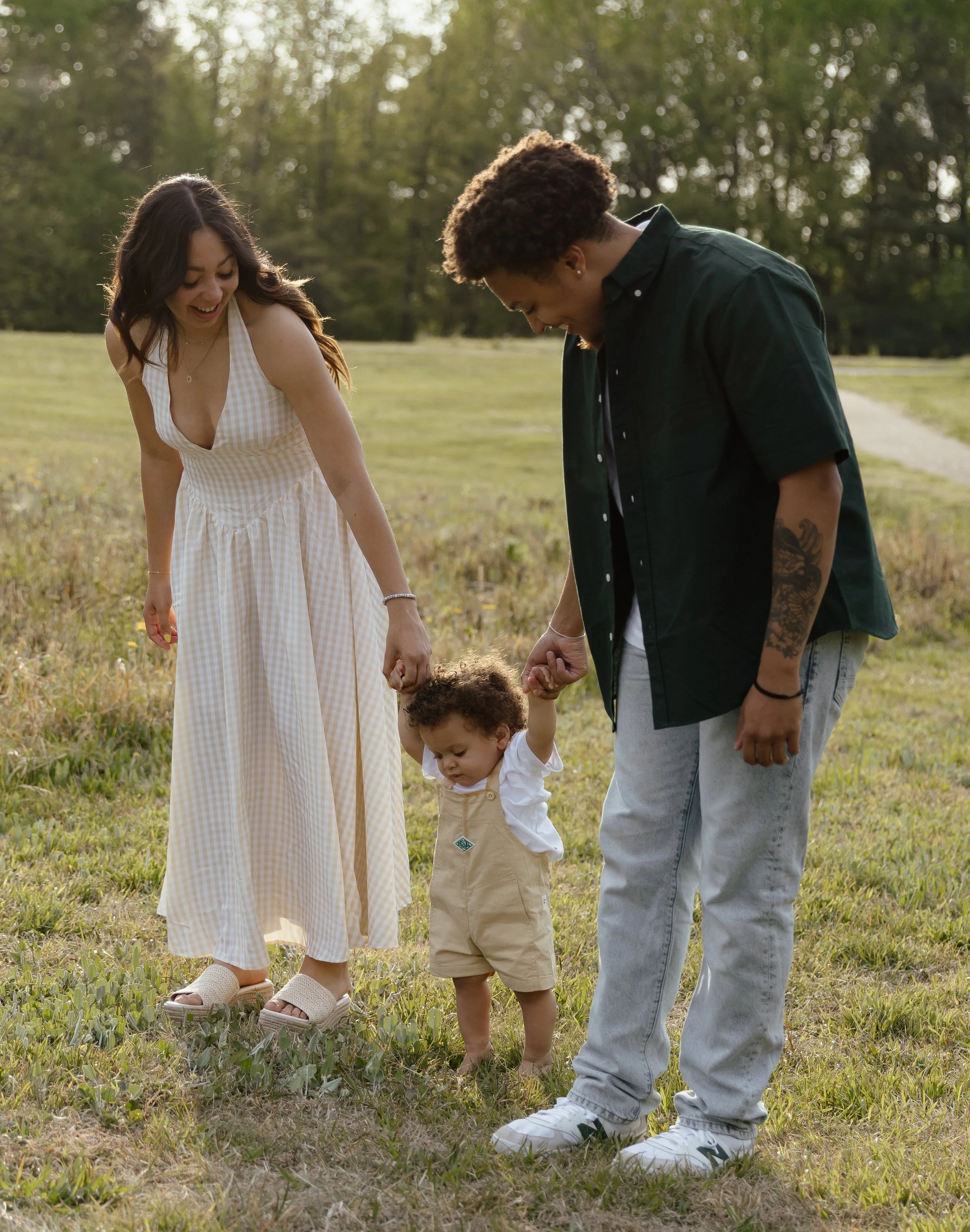 A family of three with a woman, man, and small child walking outdoors on a grassy field with trees in the background; they are holding hands and appear happy.