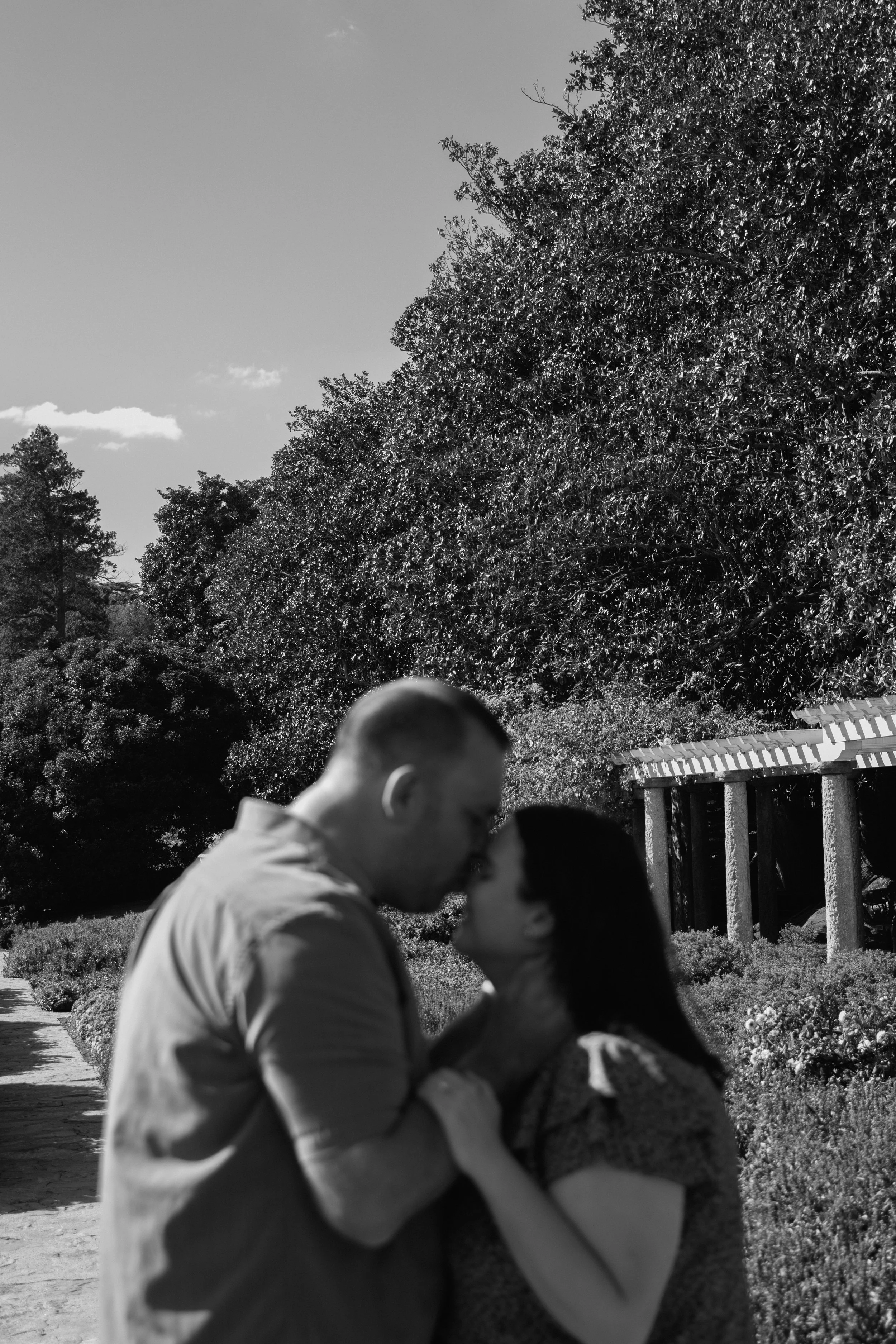 A black and white photo of a couple kissing outdoors in a park, surrounded by trees and a pathway.