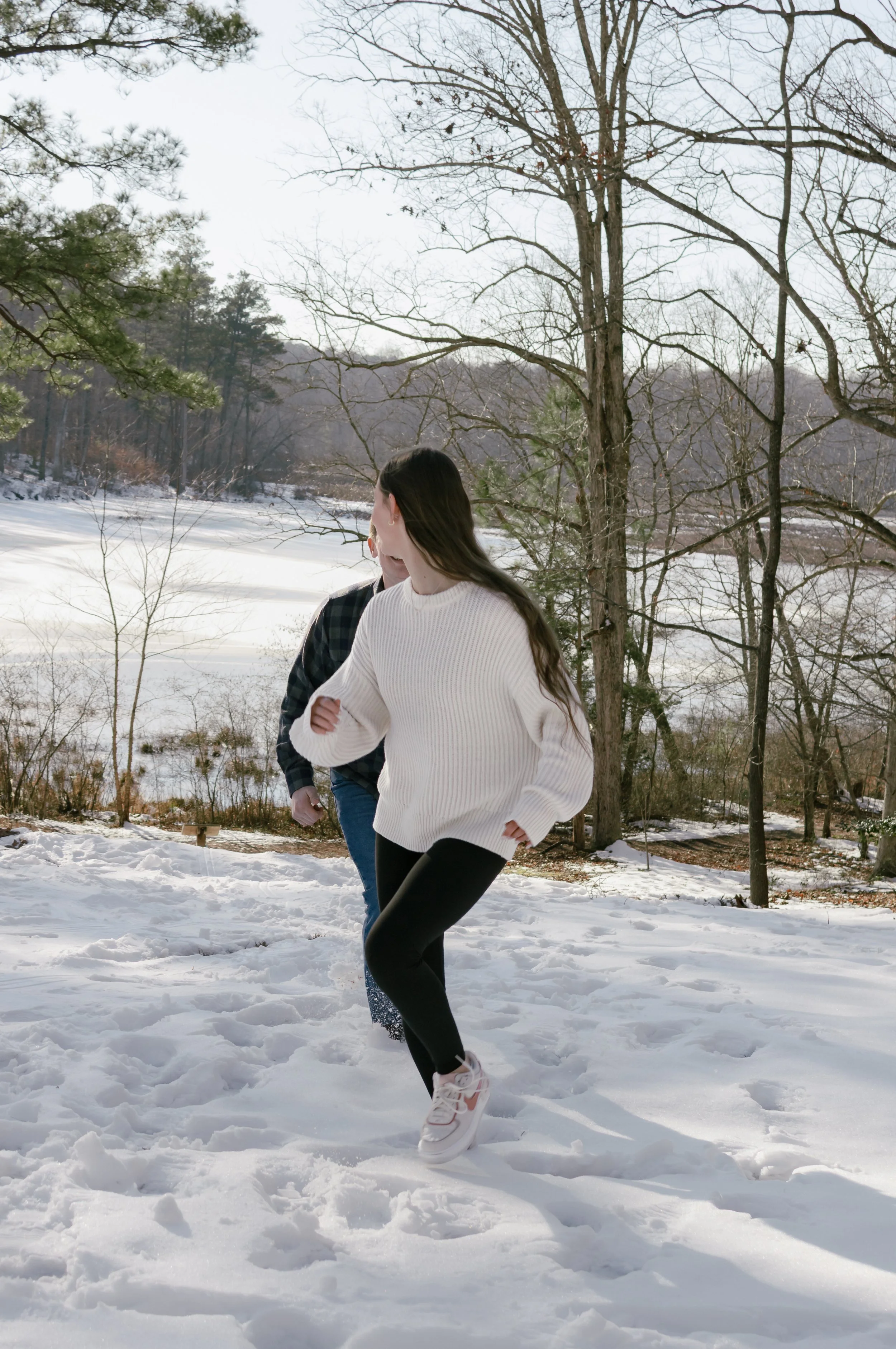 A young woman and man playing in the snow near a frozen lake with trees in the background.