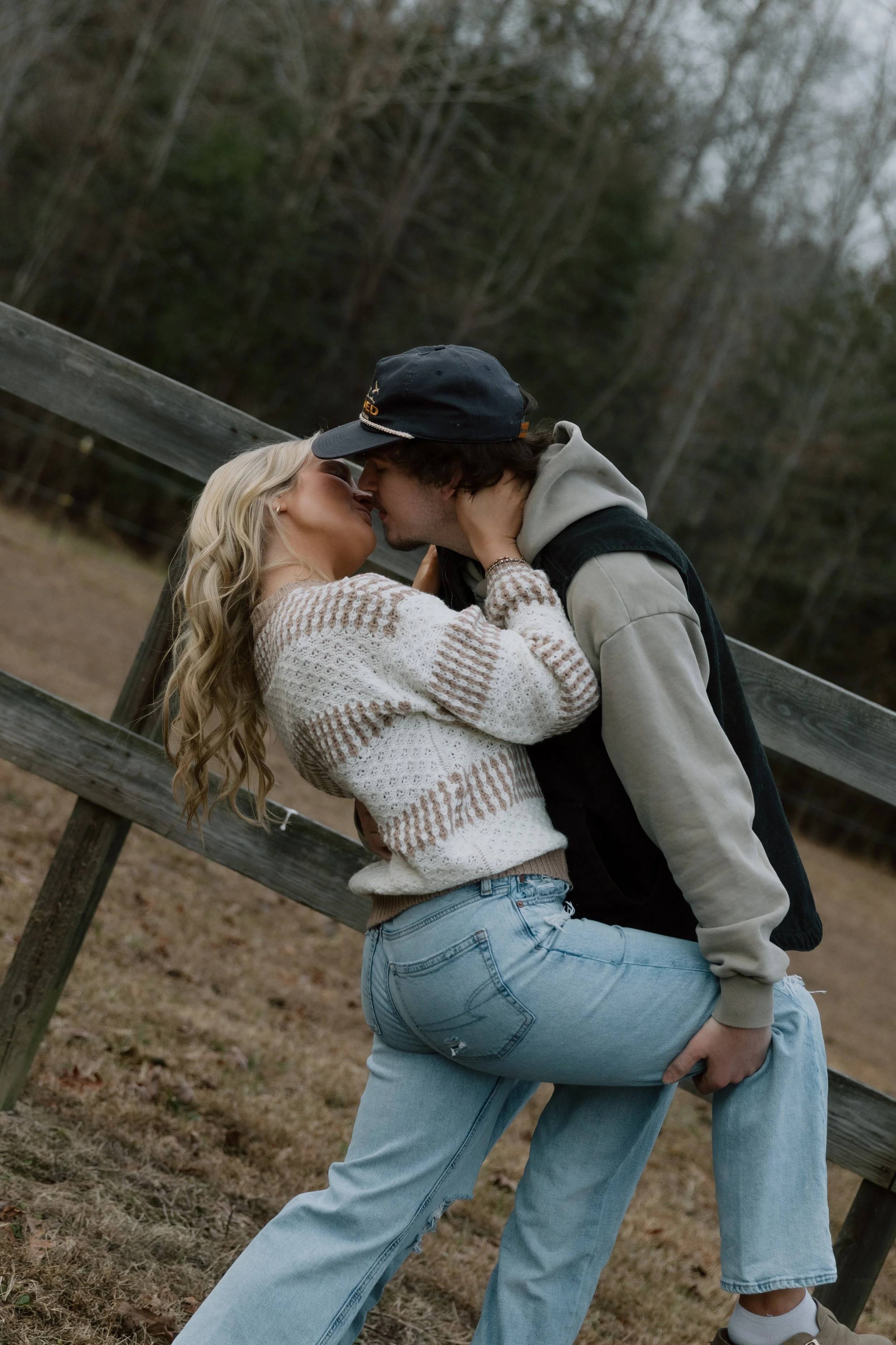 A young couple sharing a kiss outdoors near a wooden fence, with trees in the background.