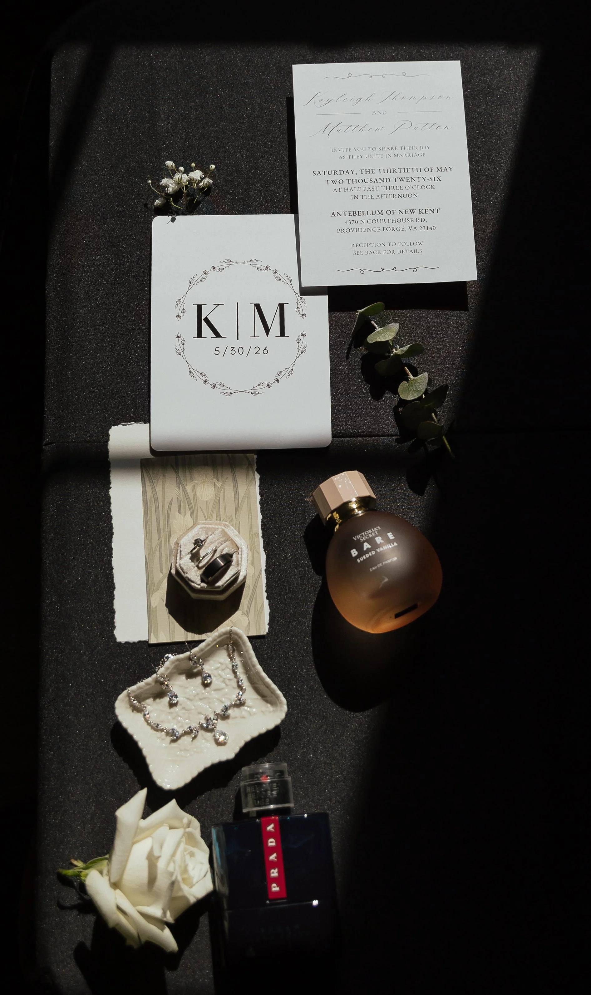 A detail photo of wedding invitations, featuring the brides perfume, the grooms cologne, loose florals of babies breathe and a rose. The flatly also includes the bride and grooms wedding rings, and the bride's necklace and earrings.