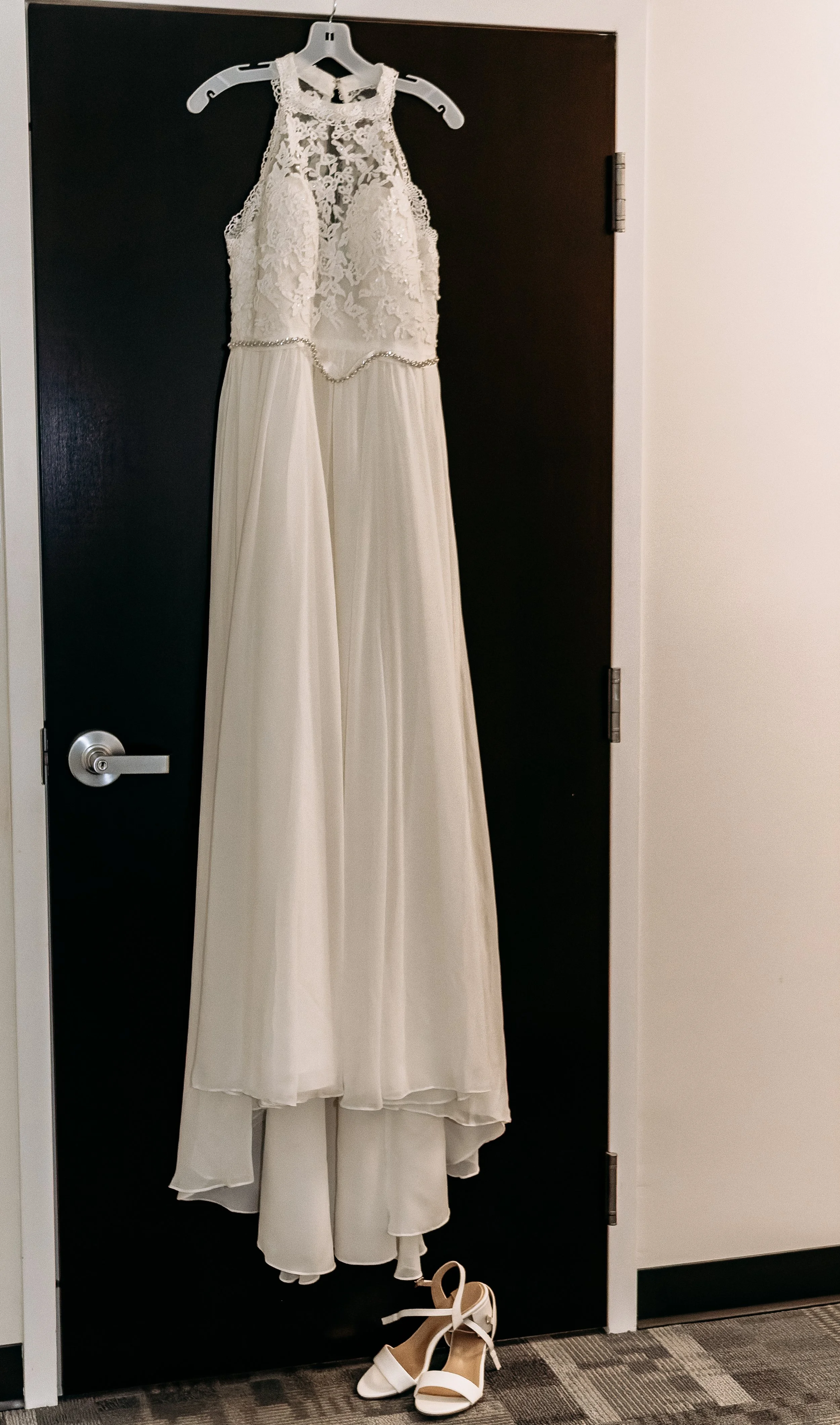 A wedding dress hanging on a black door with a pair of white heels on the floor below.