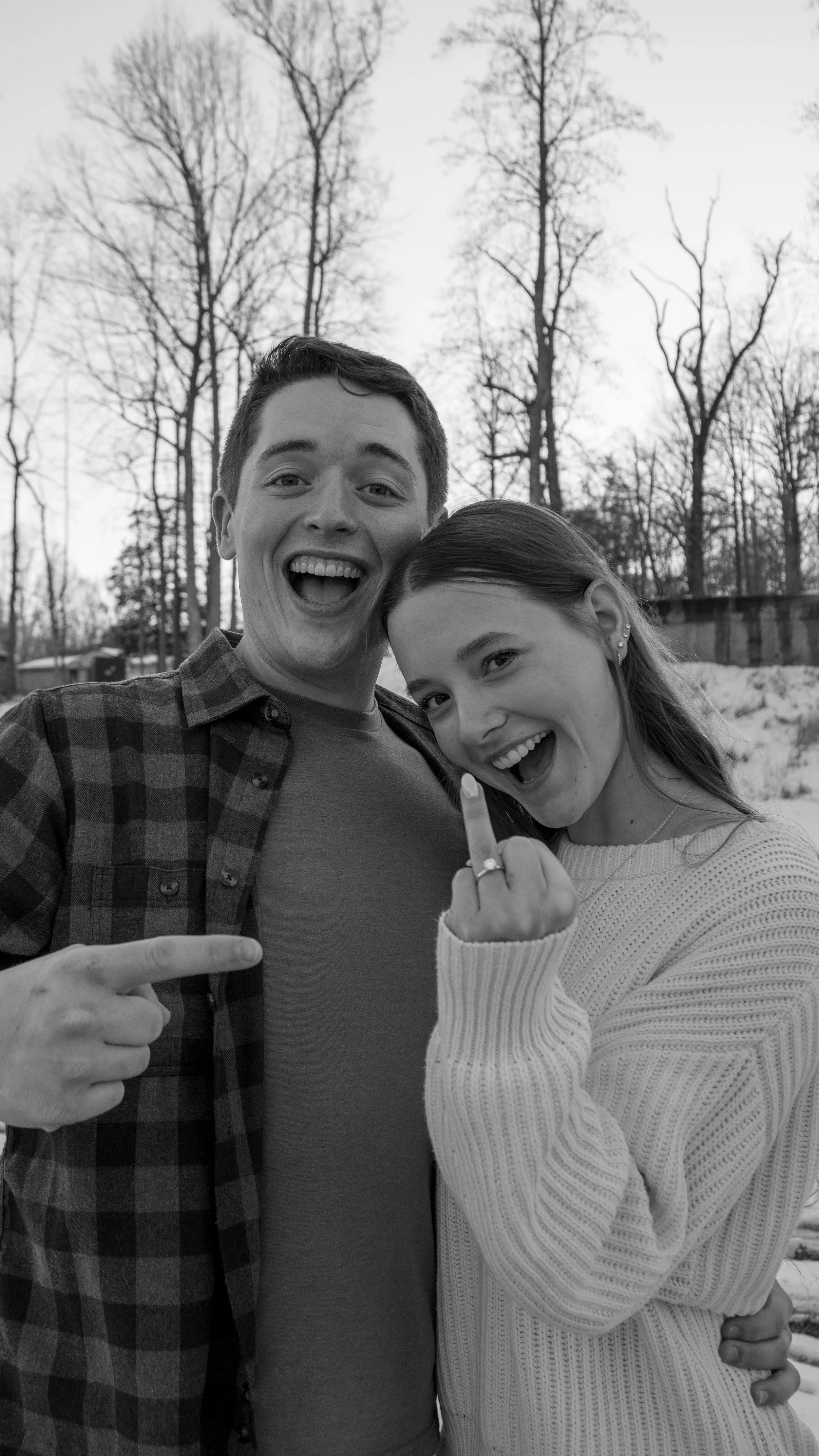 A black-and-white photo of a smiling young man and woman outdoors, with trees in the background. The woman is showing her middle finger of her right hand, which has a ring, to the camera.