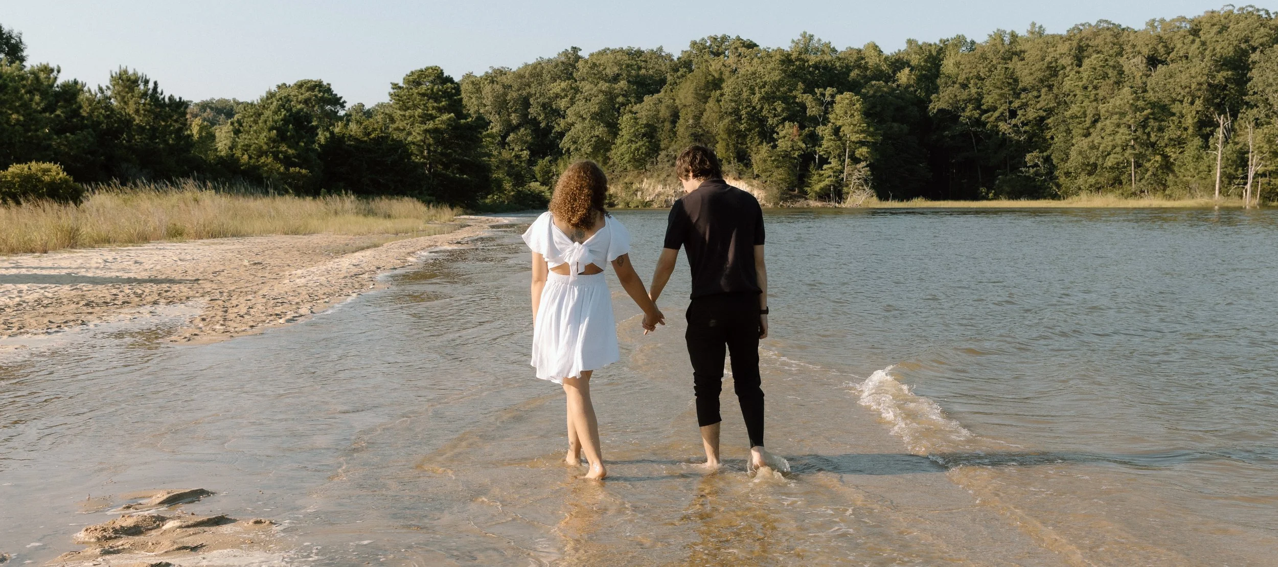 A couple holding hands, walking in shallow water along a beach with trees in the background.