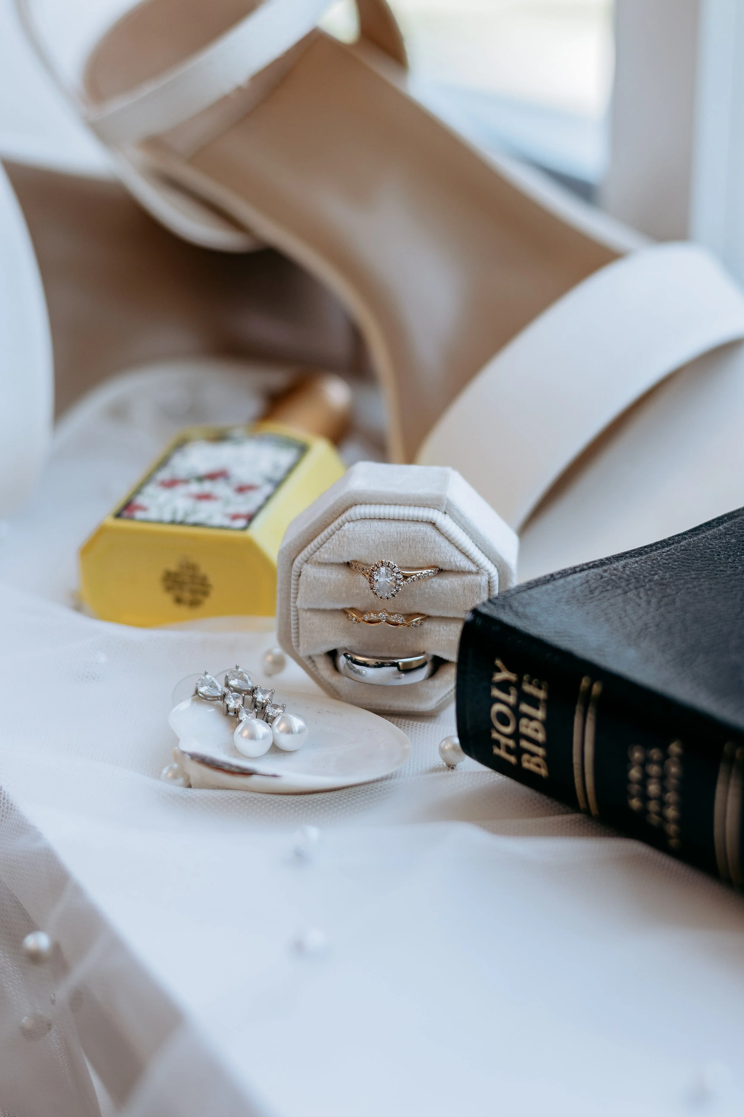 A collection of jewelry including a diamond ring, earrings, and pearl earrings, arranged around a black Holy Bible, with a pair of white shoes and a yellow perfume bottle in the background.