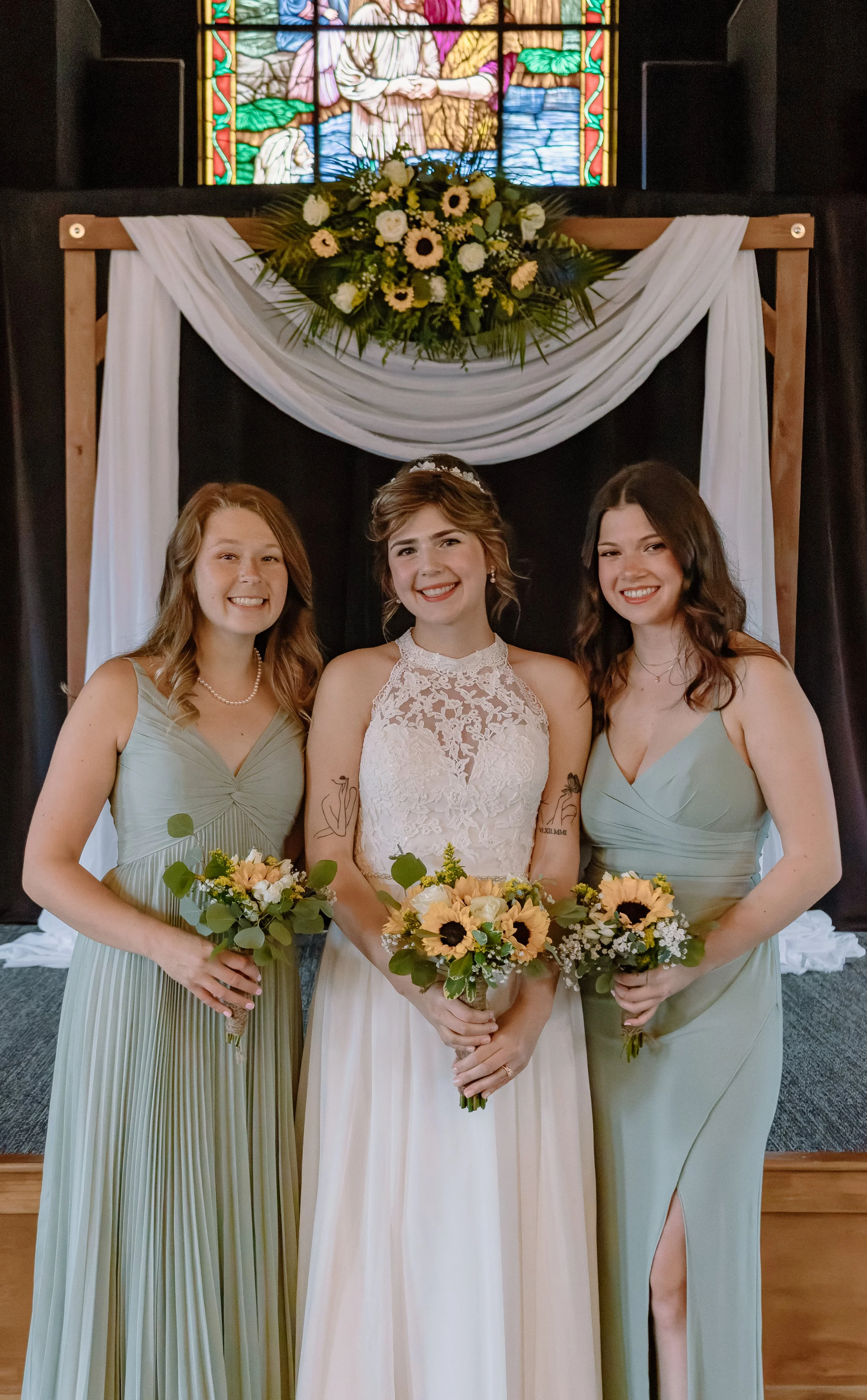 Three women standing in front of a wedding backdrop with a floral arrangement and stained glass window, holding bouquets, with the central woman dressed in a wedding gown and the other two in matching light green bridesmaid dresses.