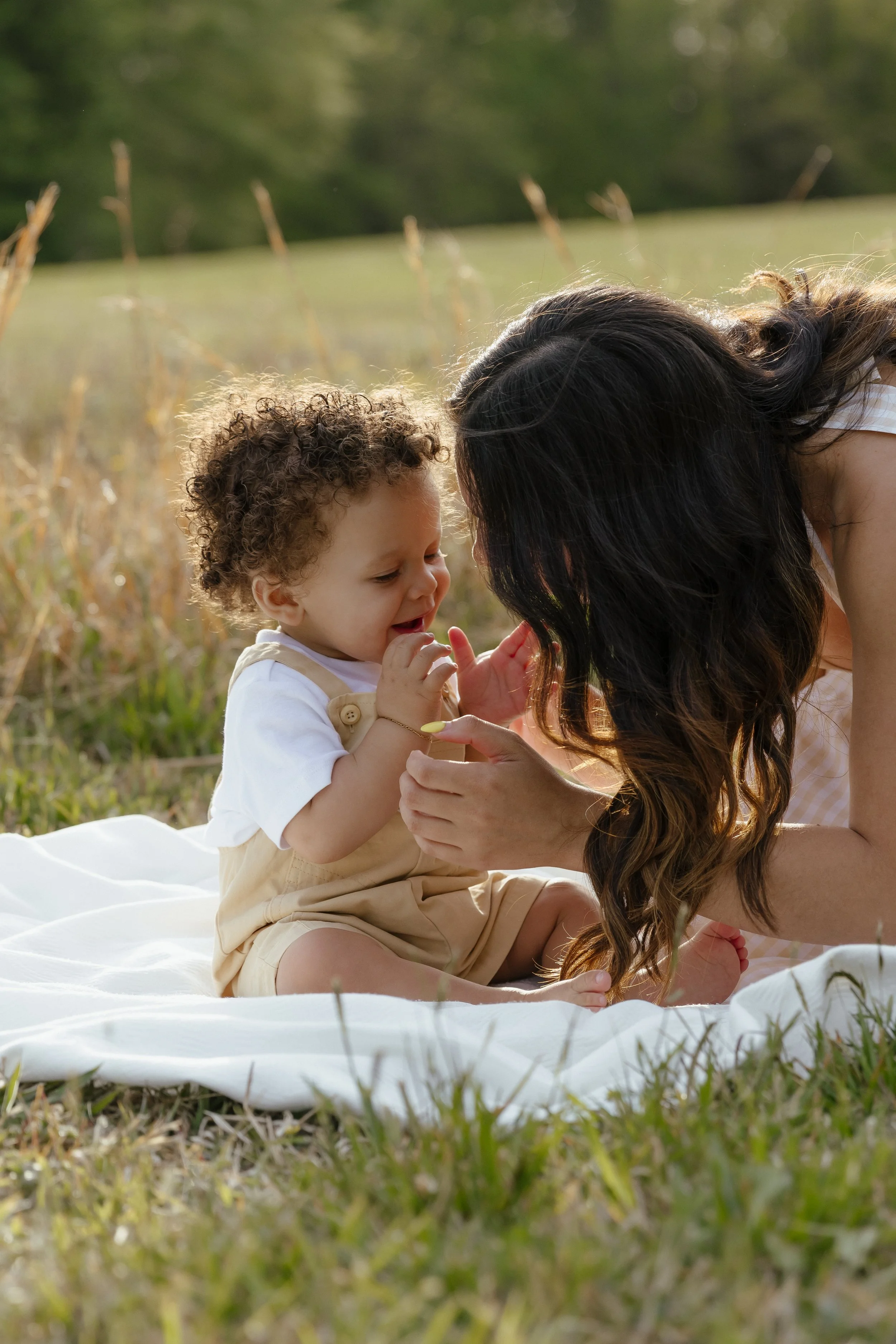 A woman and a young child playing together outdoors on a blanket in a grassy field with trees in the background.