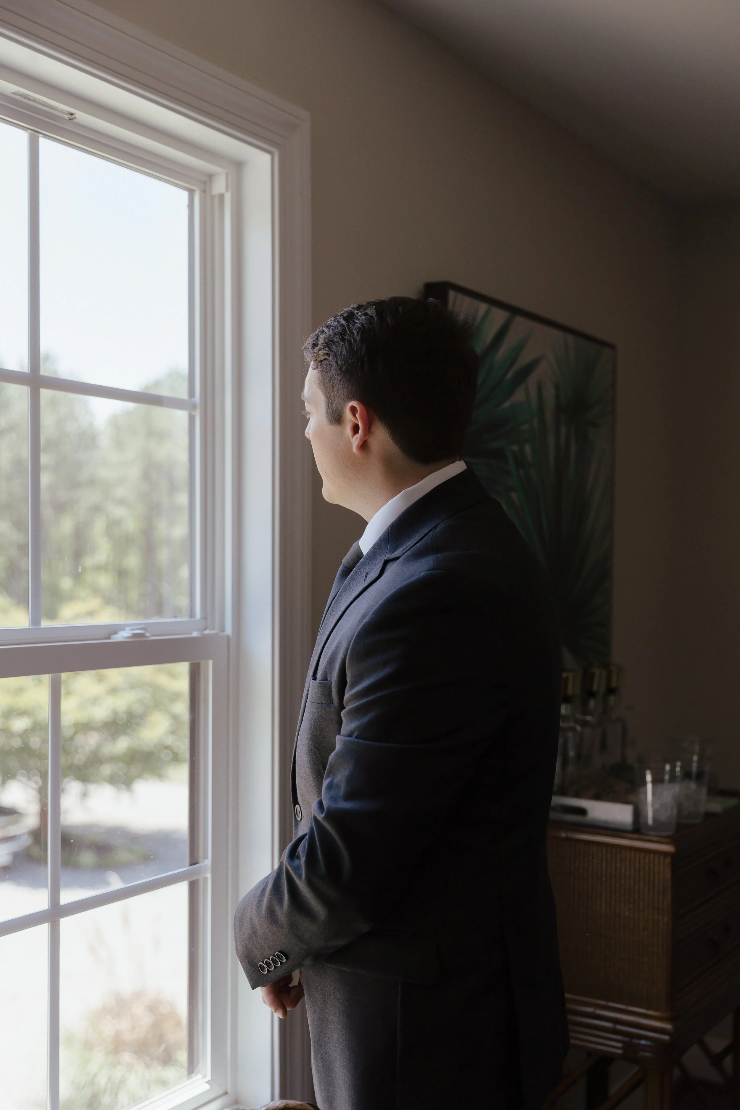 A photo of a groom who is dressed in his suit looking out the window, watching as people begin to arrive for the wedding ceremony. 