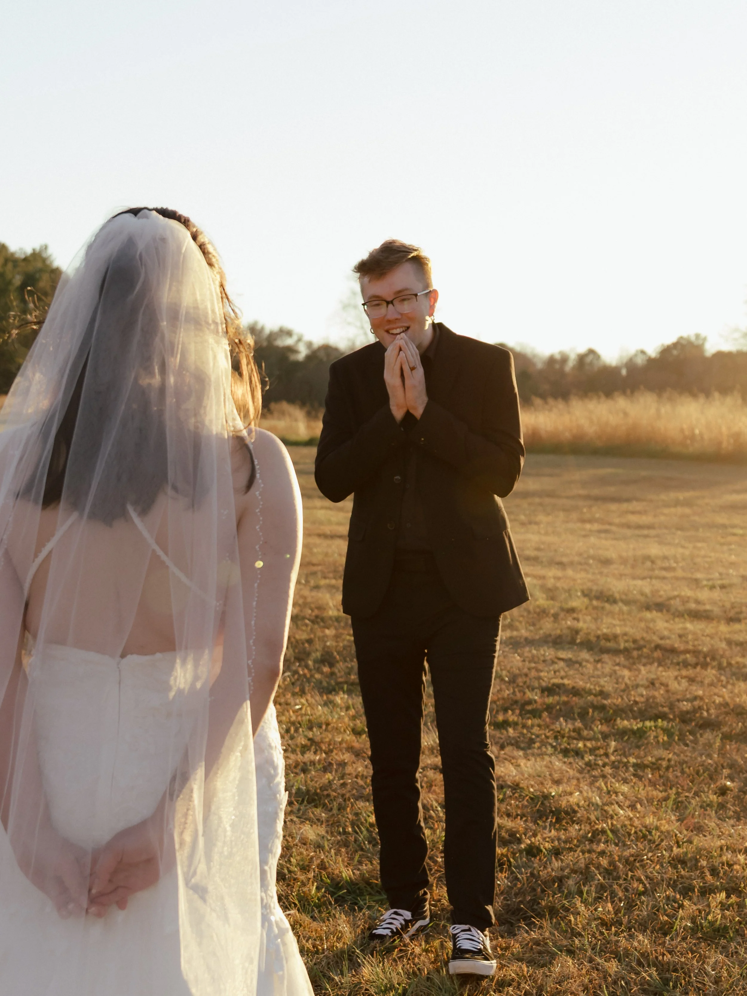 A person in a wedding dress and veil is standing outdoors, with another person in a black suit smiling with hands clasped in front of their face as the sun sets in the background.