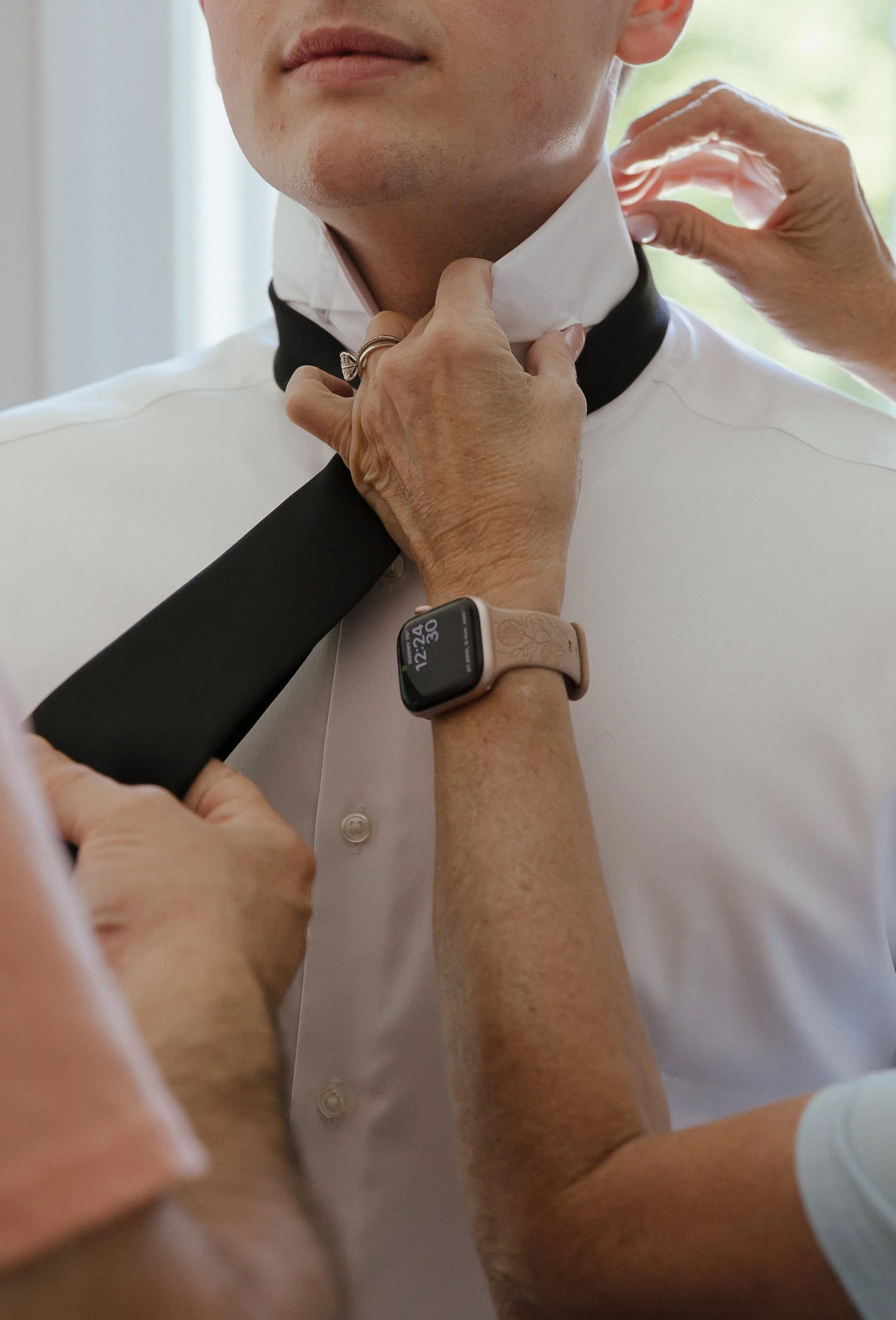 A man in a tuxedo having his collar adjusted by another person, with a blurred background and a smartwatch on the person's wrist.