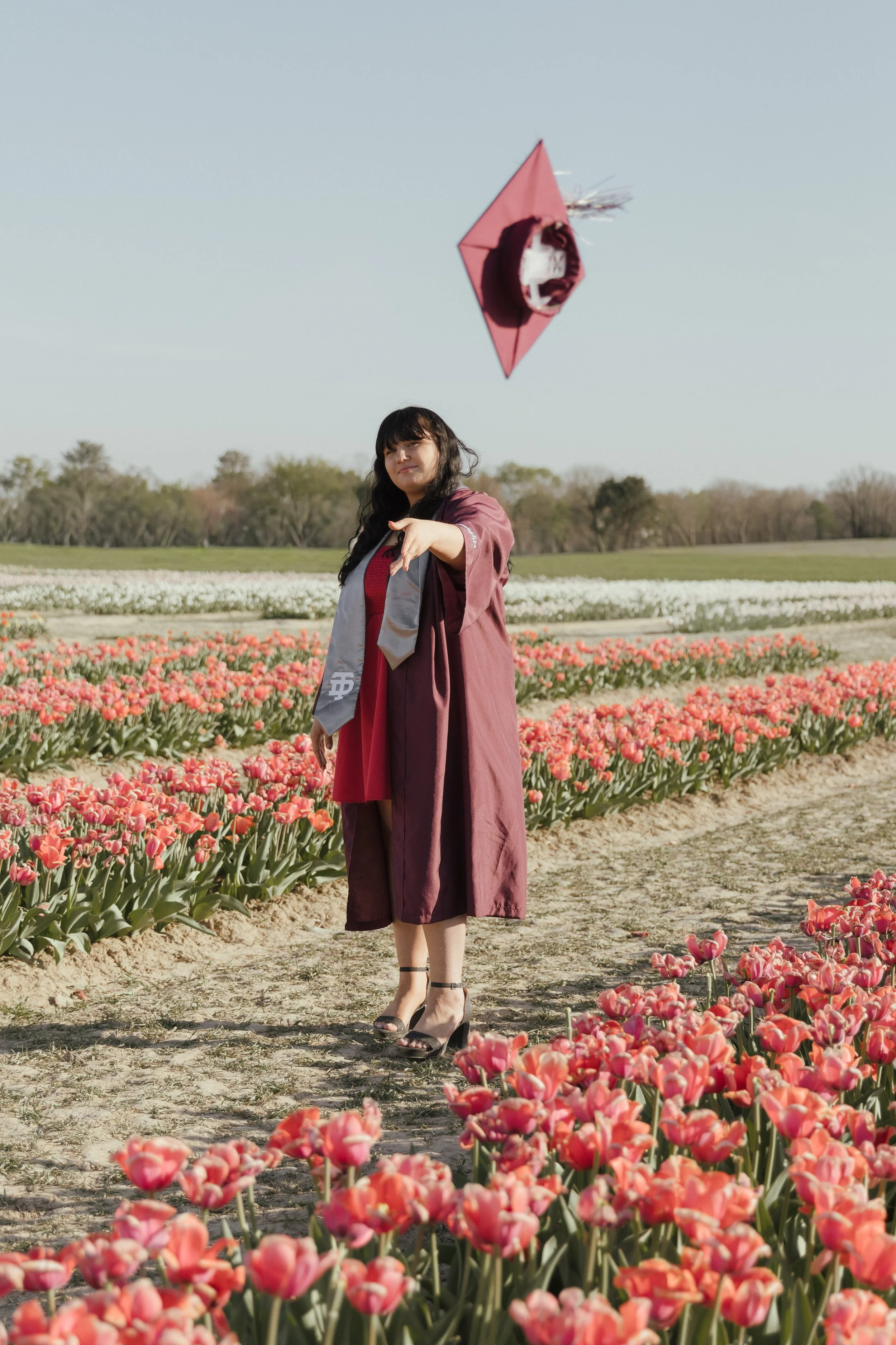 A woman in a graduation gown and cap standing on a dirt path among pink and white tulips, pointing with her finger, with a clear sky in the background.