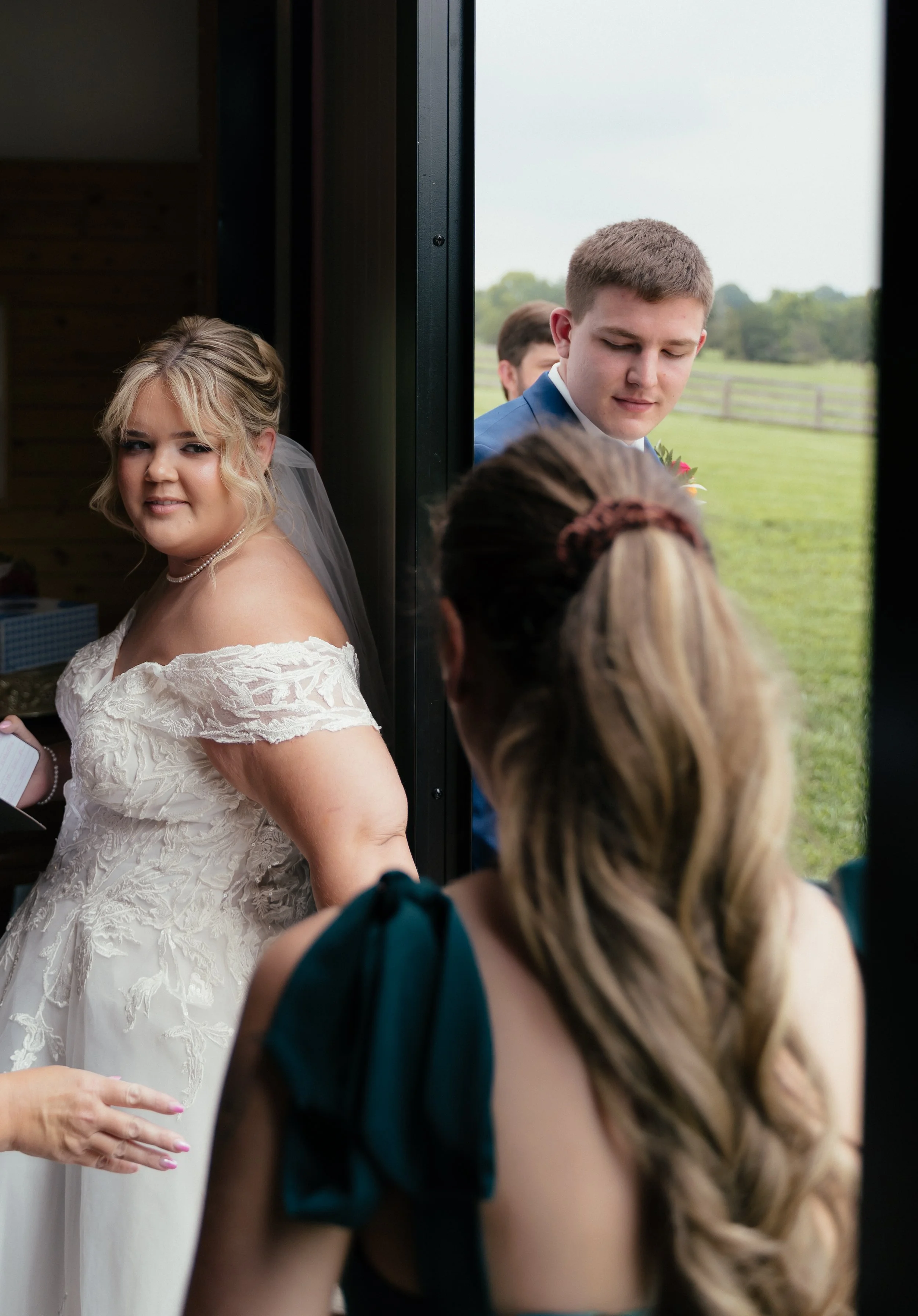A photo of a bride and groom during a first touch moment. A woman is facing them and speaking with them while standing on either side of a barn door.