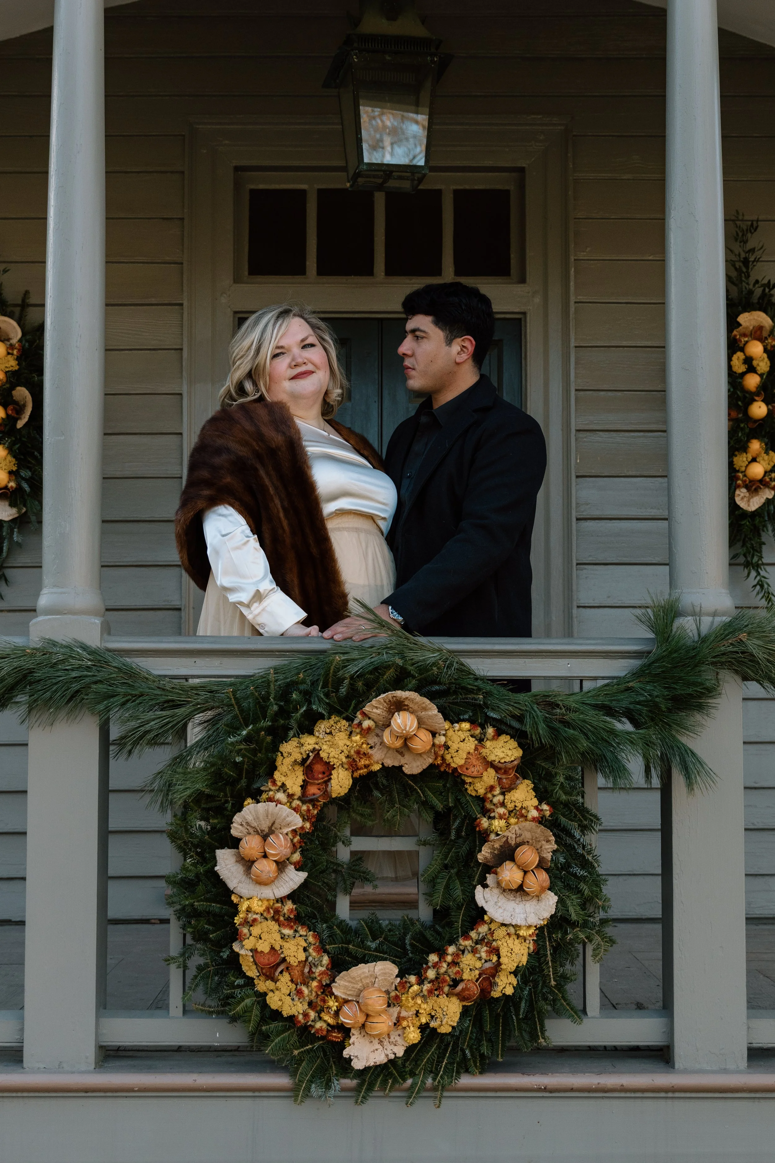 A couple standing on a decorated porch, with the woman wearing a fur stole and the man in a dark jacket. The porch has a festive wreath with yellow flowers and natural elements.