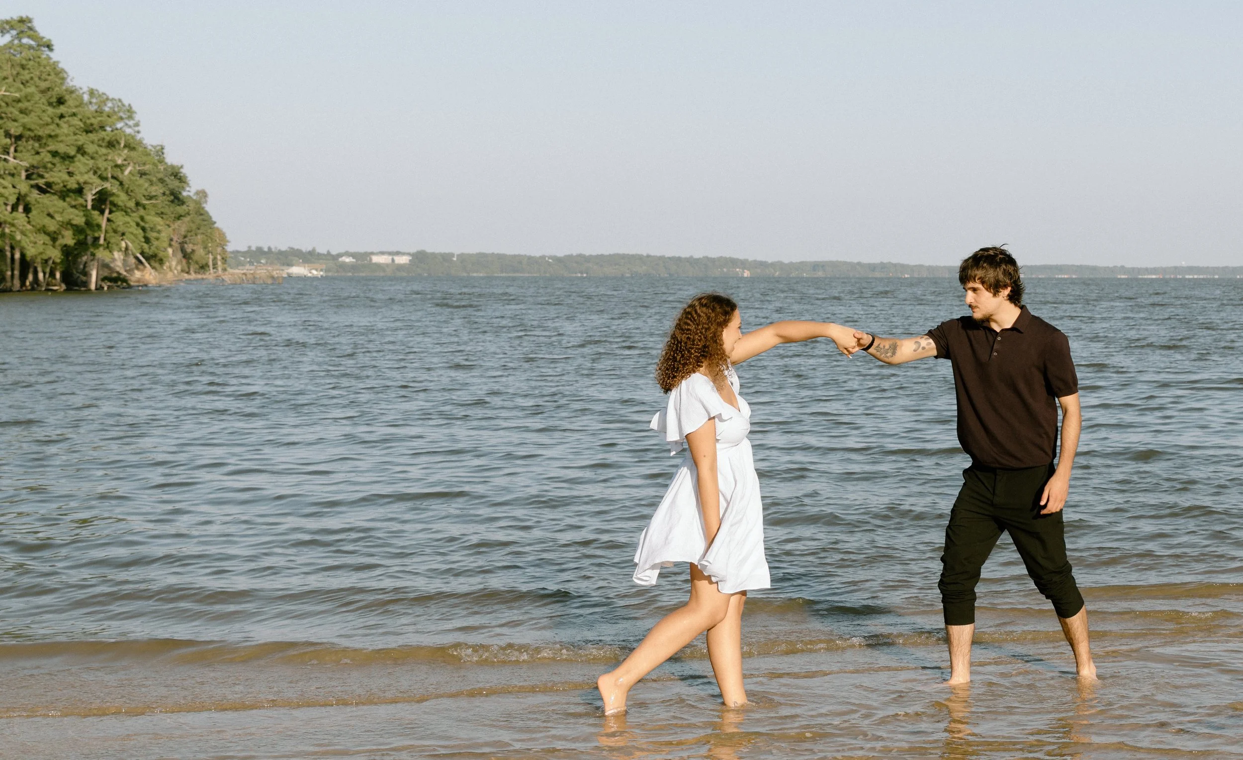 A couple holding hands and reaching out to each other on a beach near the water, with hills and trees in the background.