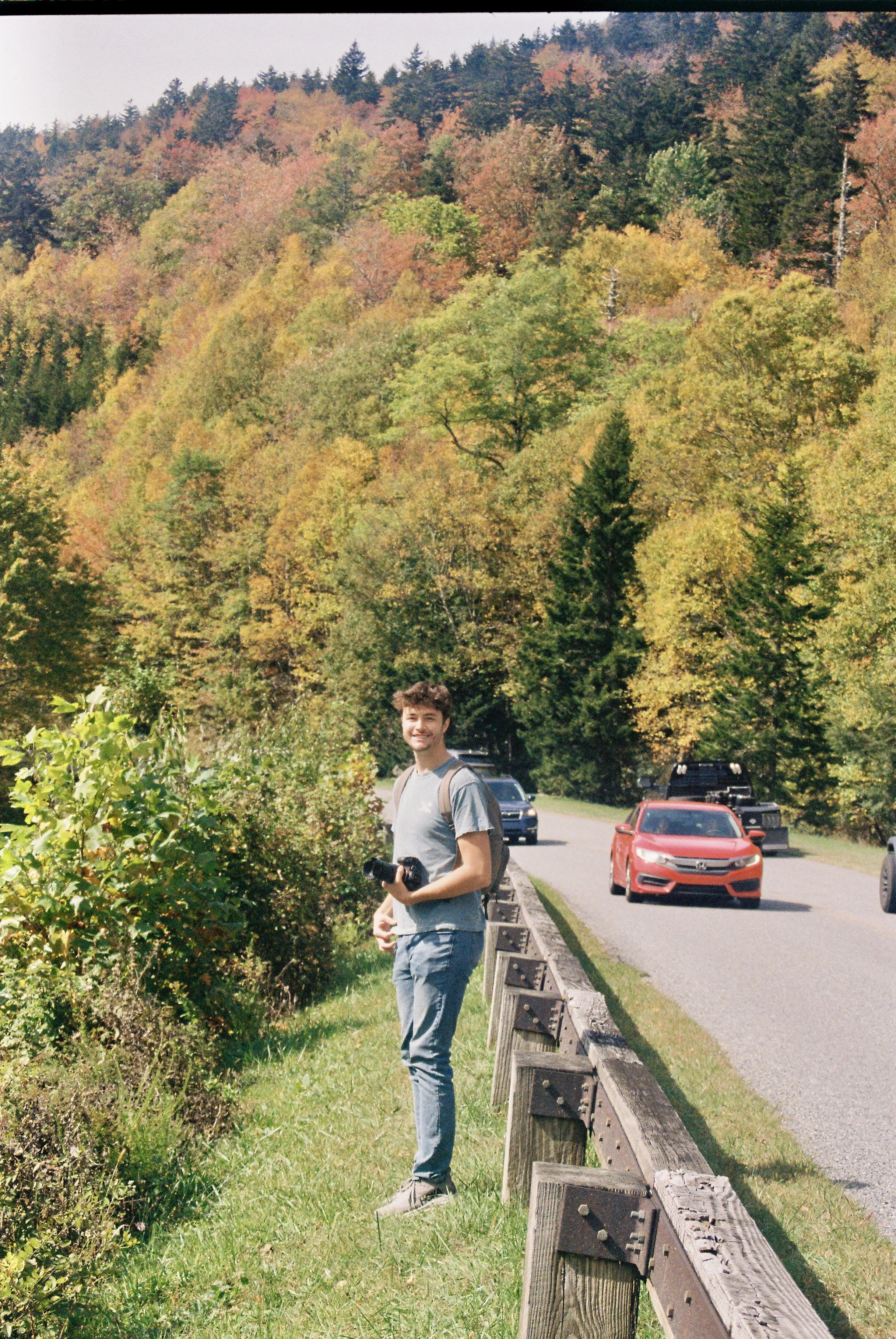 A young man standing by the roadside holding a camera, with a background of colorful autumn trees and passing cars.