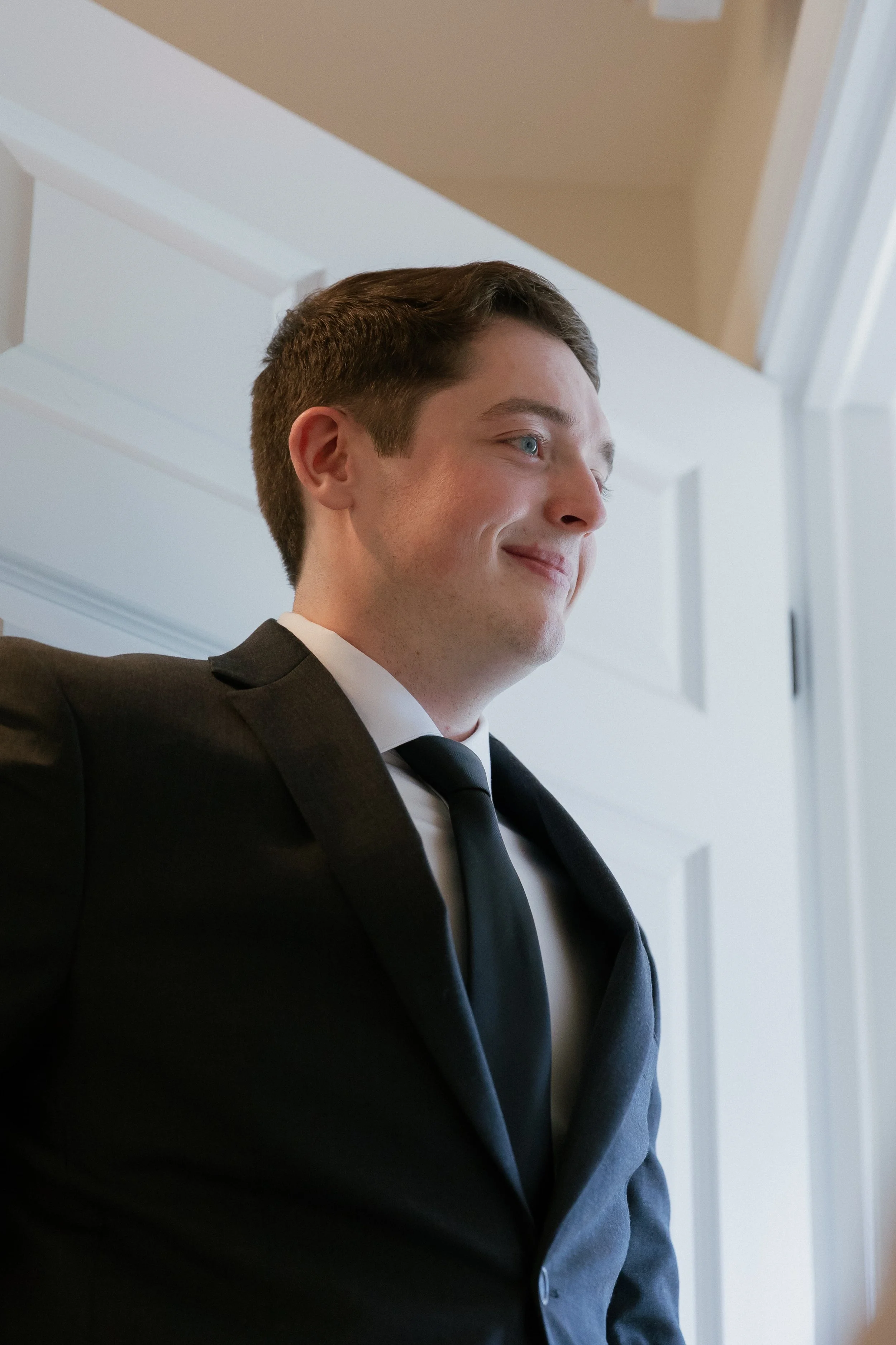 Close-up of a young man with brown hair, blue eyes, wearing a black suit and white shirt, smiling gently, standing indoors near a white door.