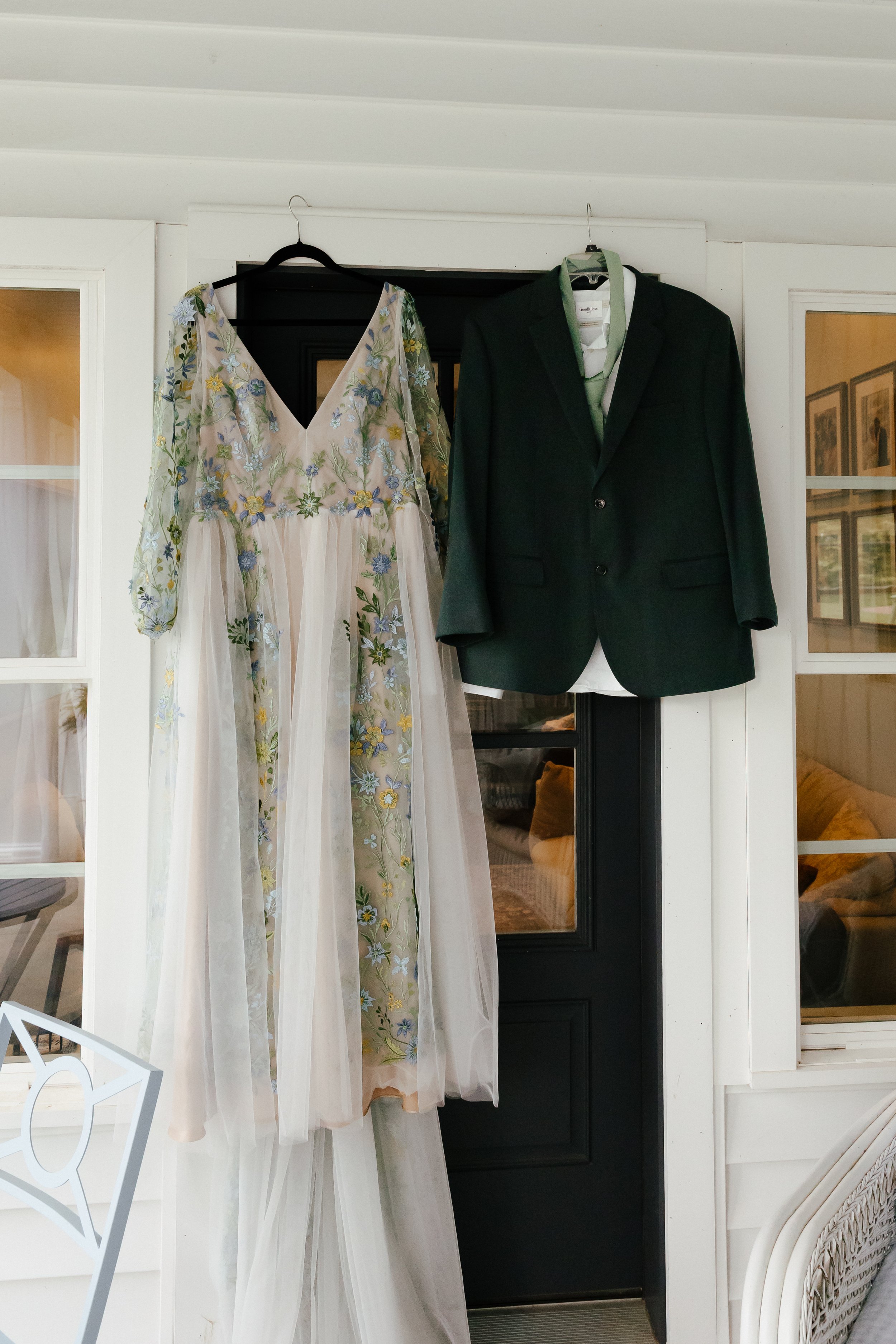 A photo of a wedding dress with lace colorful floral details hanging outside of a door next to a dark green suit with a sage green tie.