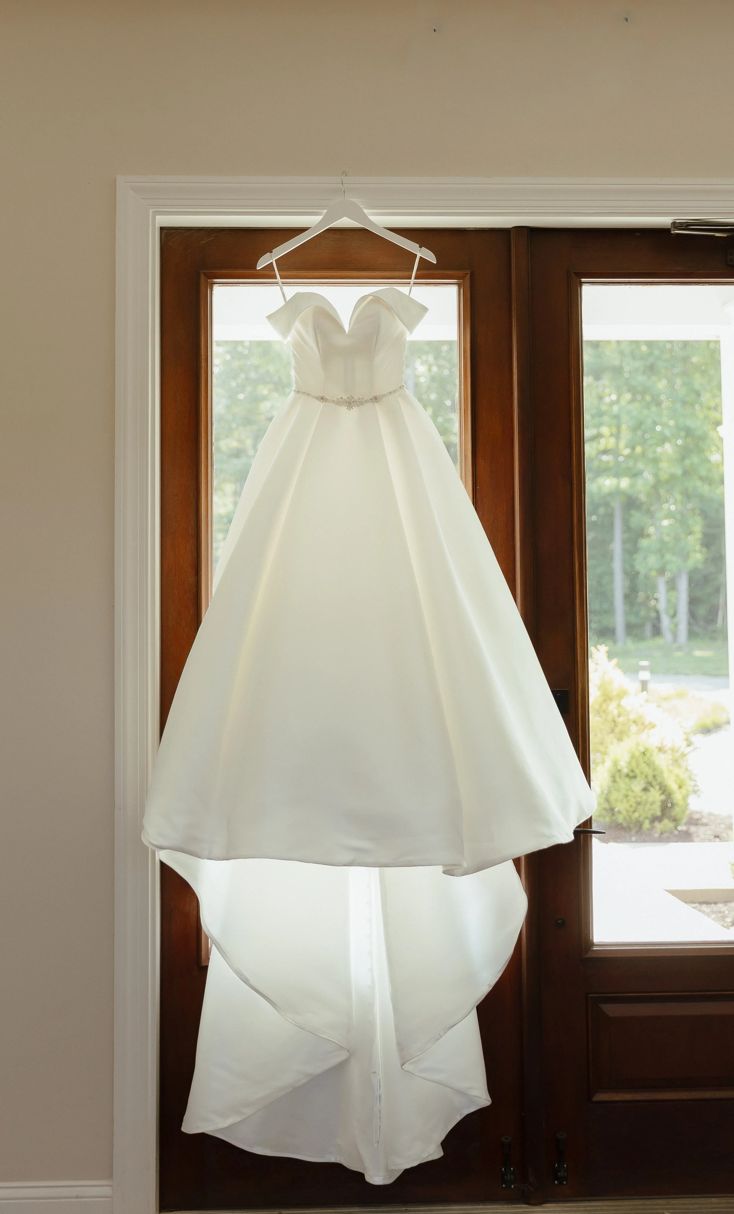 A photo of a wedding gown hanging against a wooden glass door. The wedding dress is an off the shoulder ballgown, and has a simple and elegant jeweled belt.