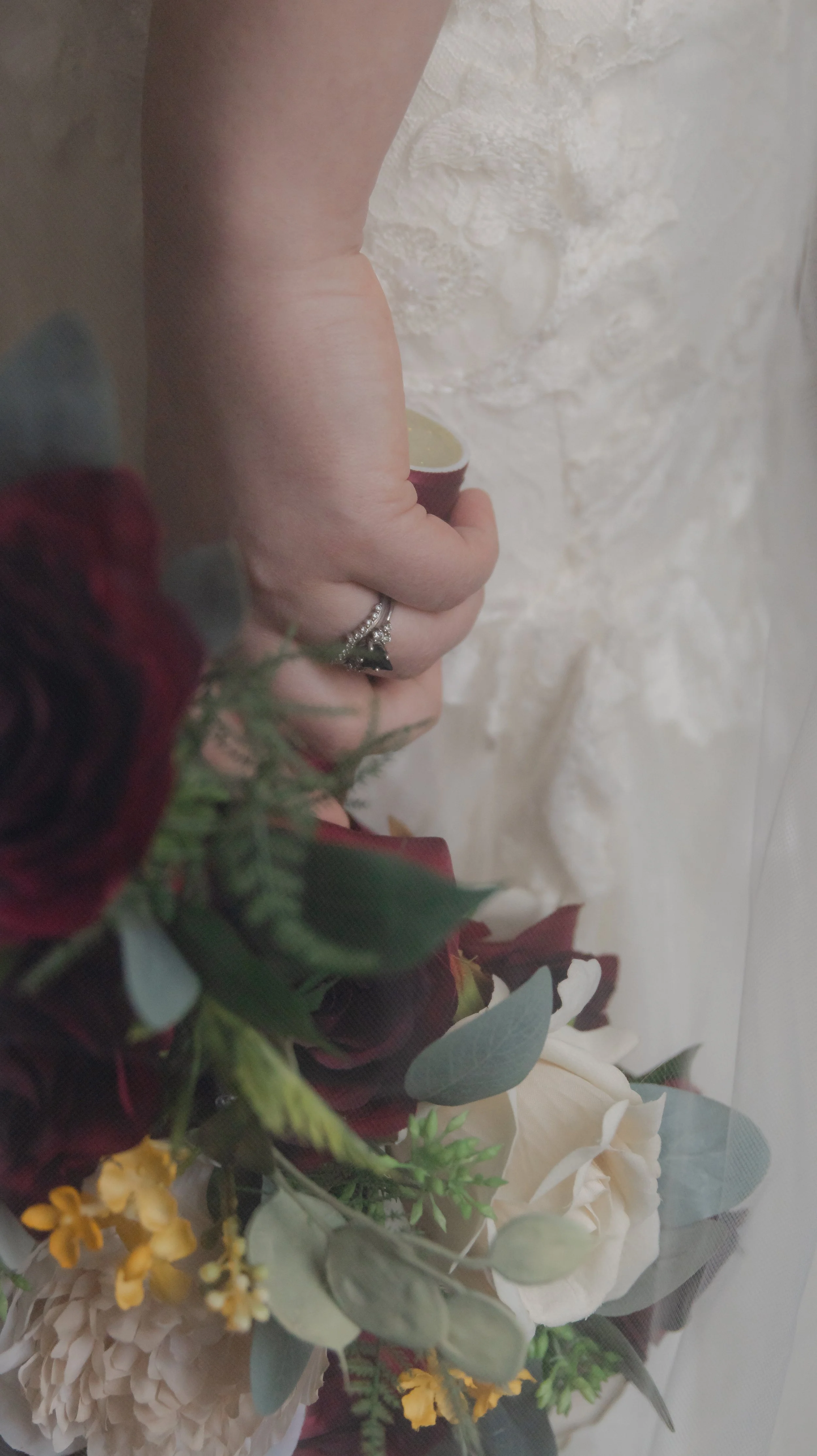 Close-up of a hand with a wedding ring holding a small bouquet of flowers against a textured cream-colored wall.