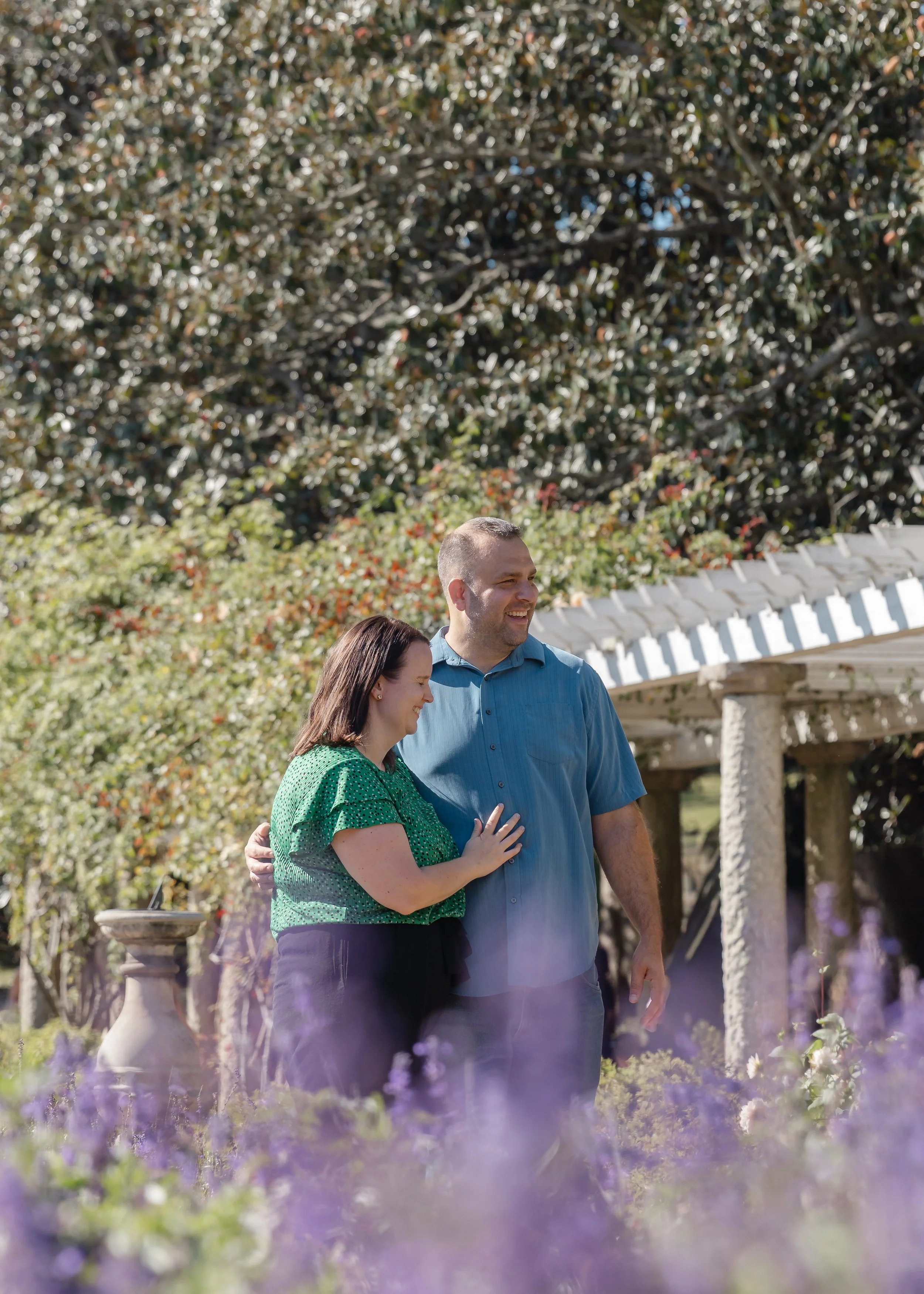 A smiling couple standing together outdoors in a garden, with greenery and a small white gazebo in the background.