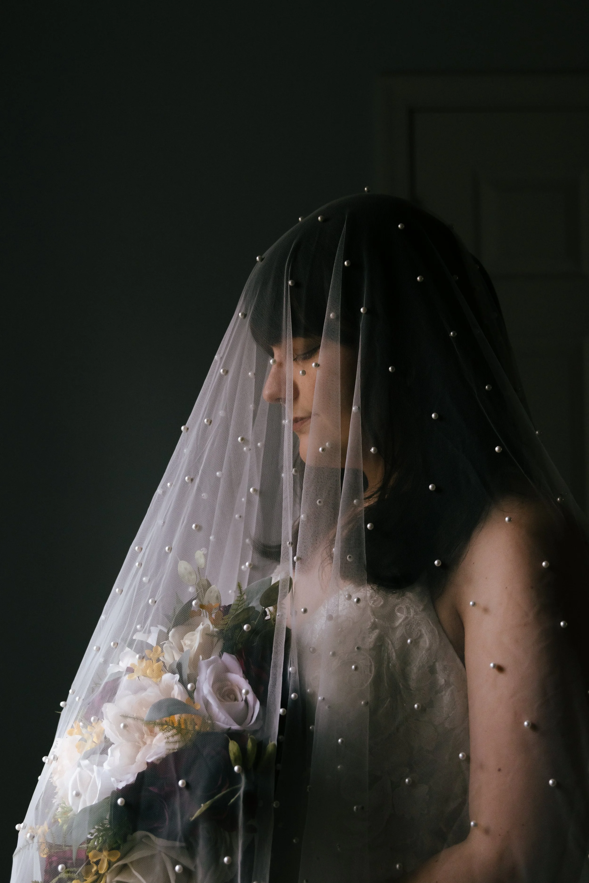 A woman with dark hair and freckles wearing a wedding veil decorated with pearls, holding a bouquet of flowers, against a dark background.