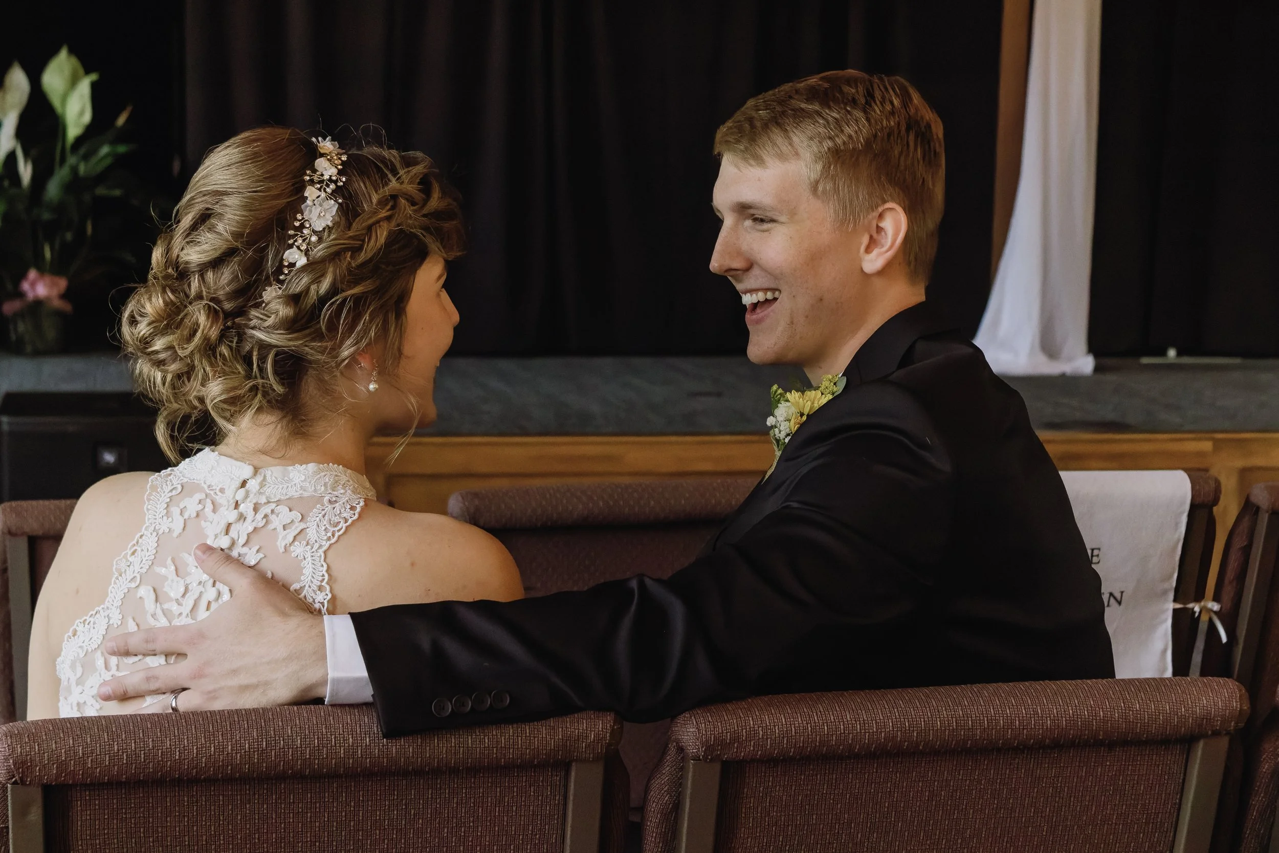 A bride and groom sitting close together during their wedding ceremony, smiling and embracing each other inside a decorated venue.