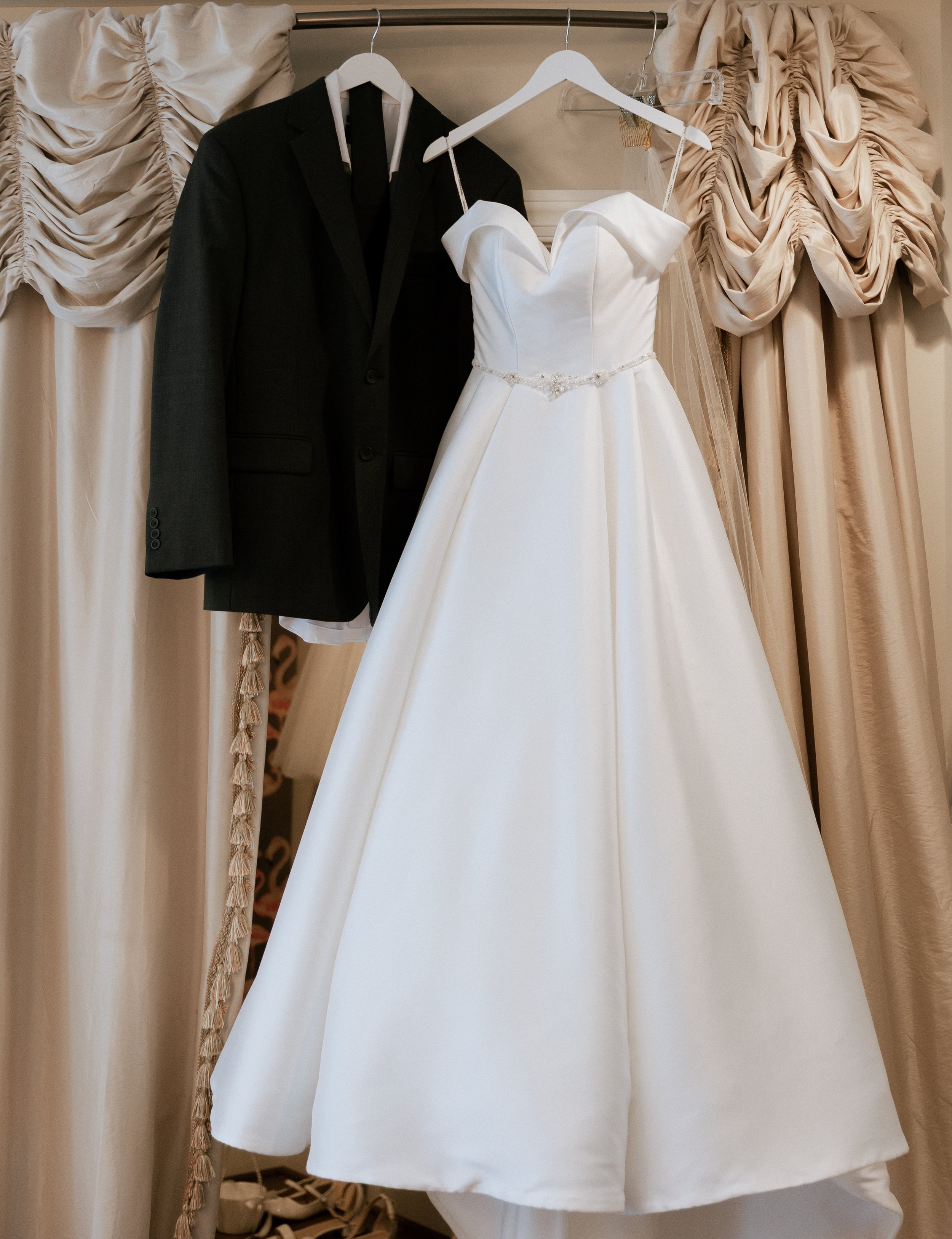 A wedding dress with a sweetheart neckline and a beaded belt on a hanger, alongside a black suit jacket on a mannequin, hanging in front of cream-colored, ruched curtains.