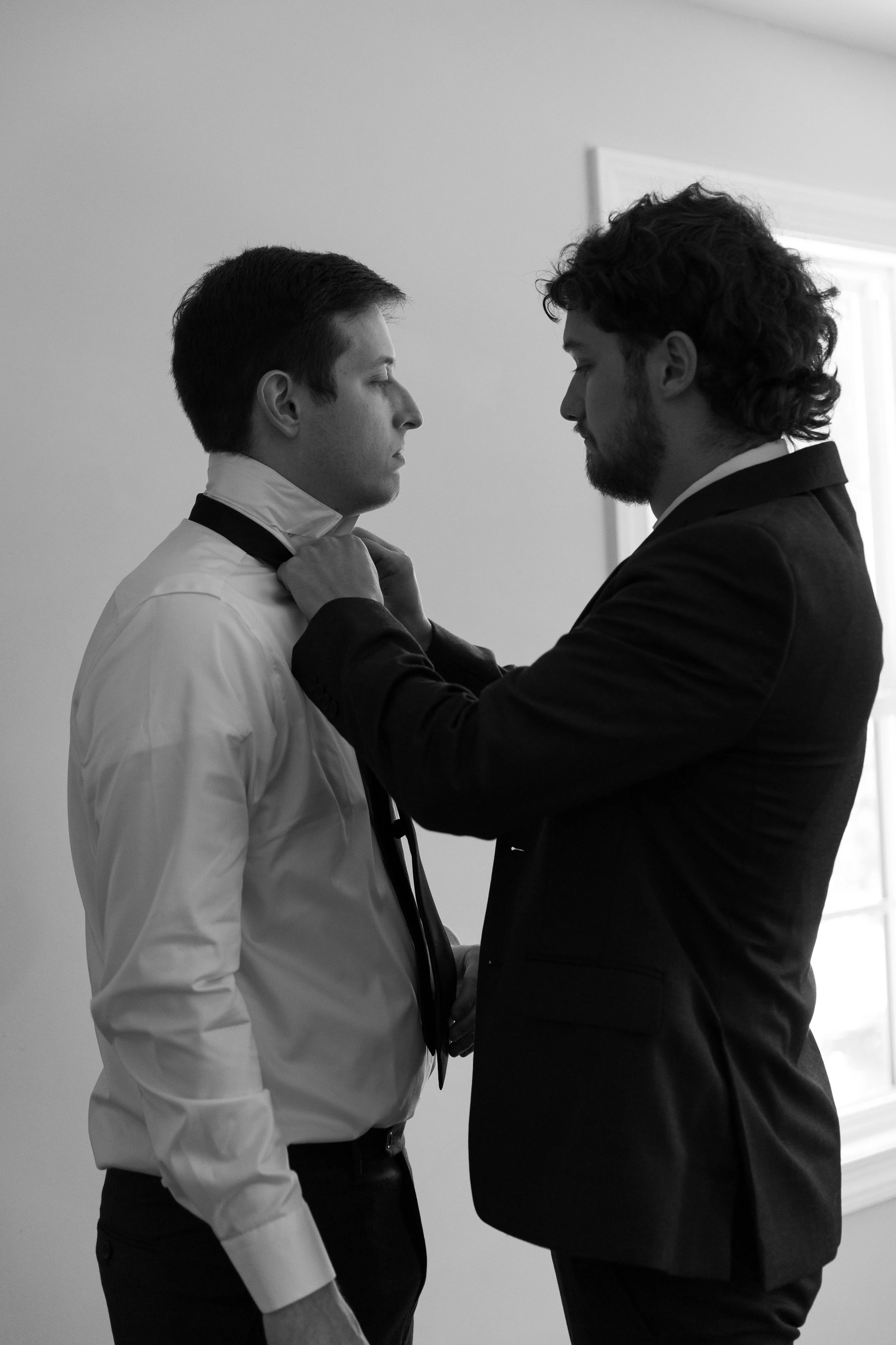 Two men, one in a white shirt and the other in a dark suit, face each other indoors as they prepare to tie a tie, with the man in the dark suit helping the other.