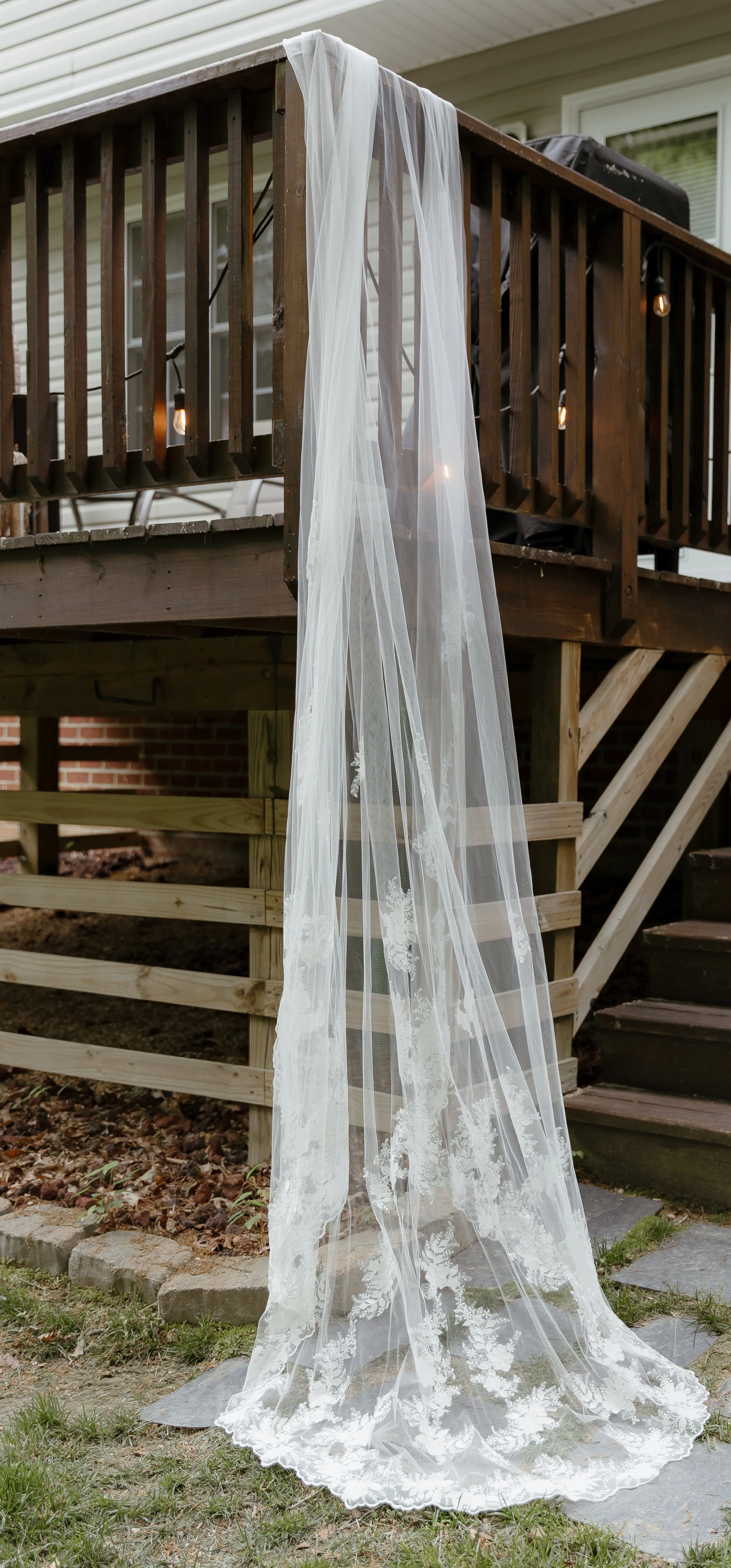 A sheer white wedding veil with floral embroidery hanging from a wooden deck railing outside a house.