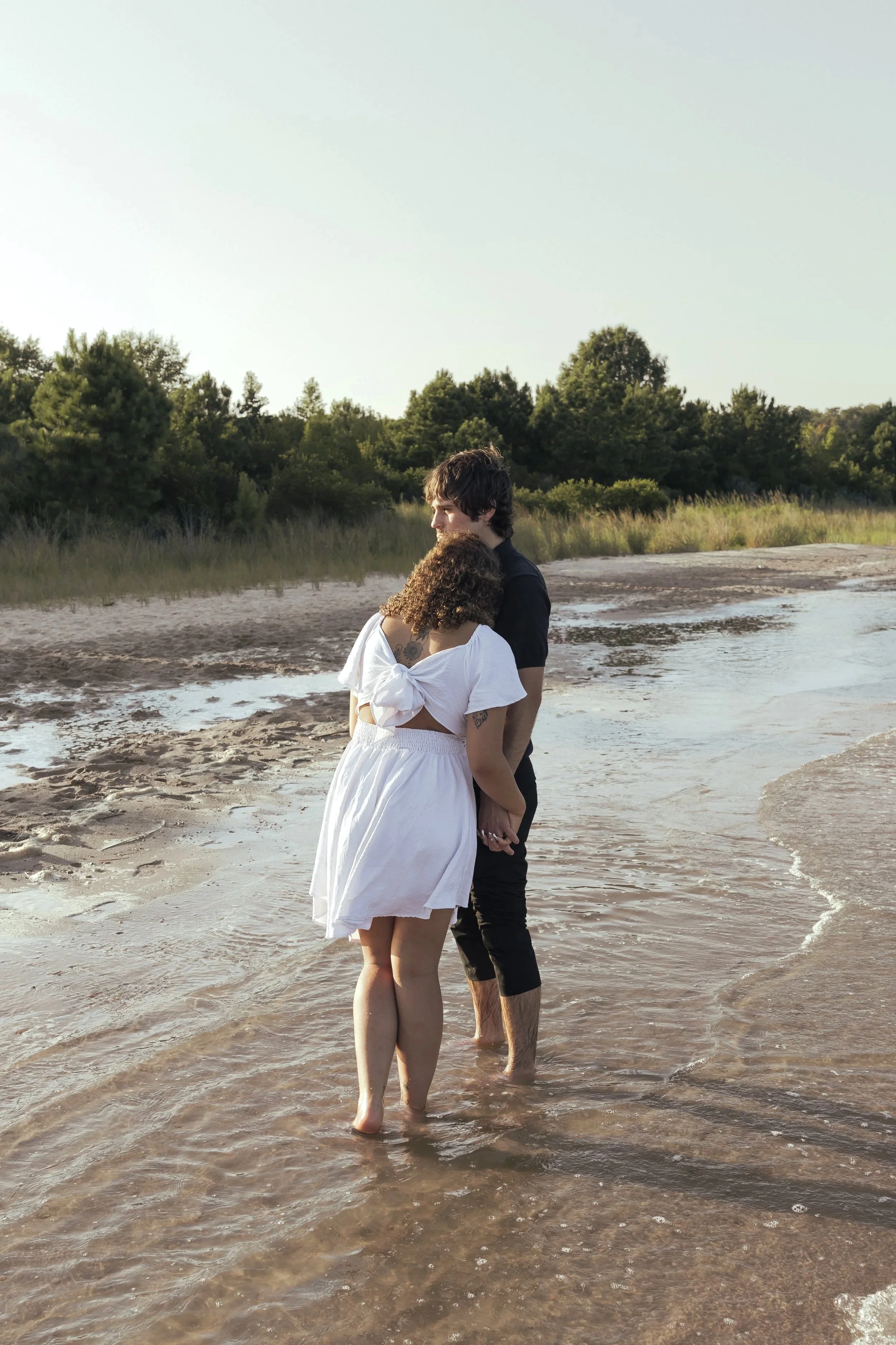 A couple standing together on the beach with water around their ankles, holding hands and embracing, during sunset with trees in the background.