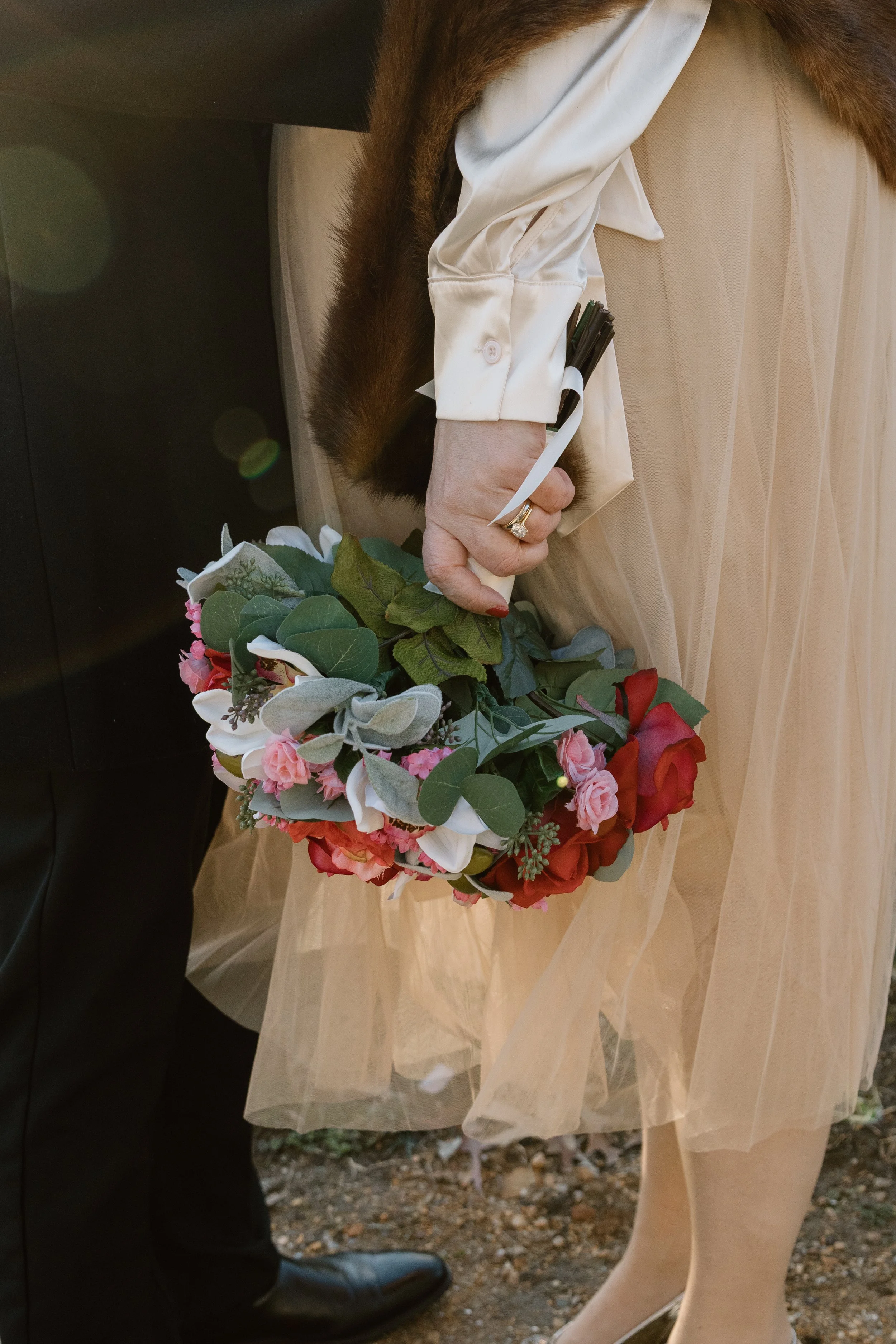 A person in a beige dress and white stocking holds a colorful bouquet of flowers, standing next to a person in black formal shoes.