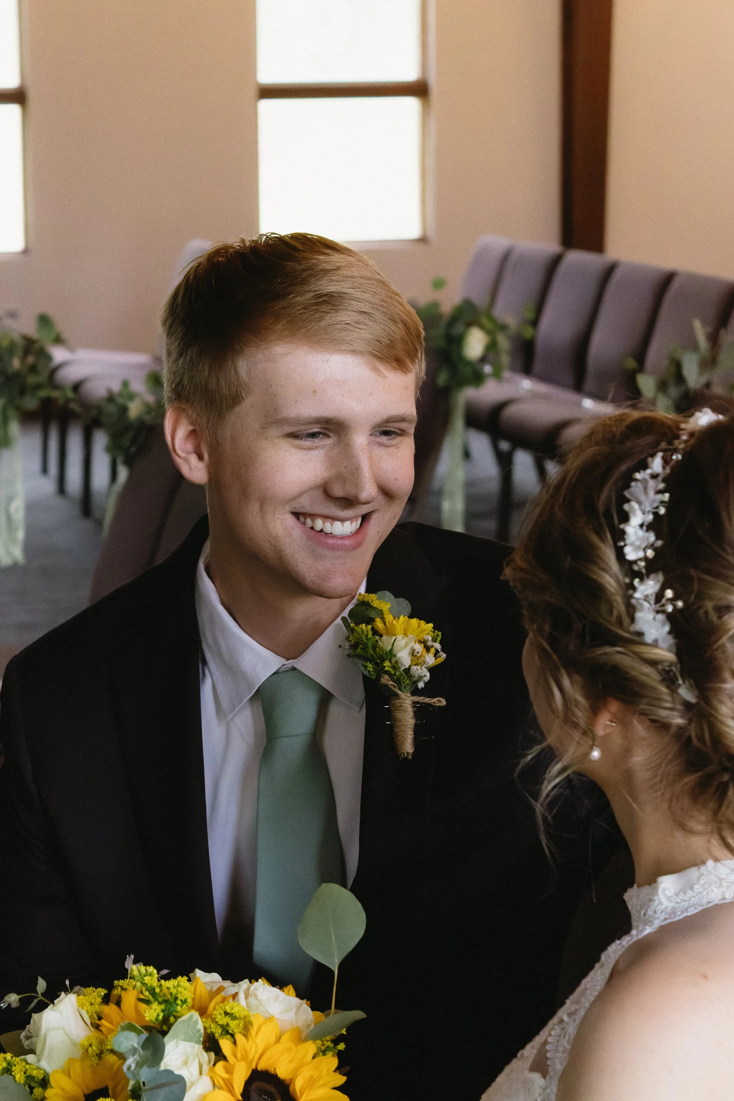 A young groom with a boutonniere smiles during a wedding ceremony, sitting across from the bride, who is wearing a white lace dress and a floral hairpiece. The venue has rows of chairs decorated with flowers and greenery.