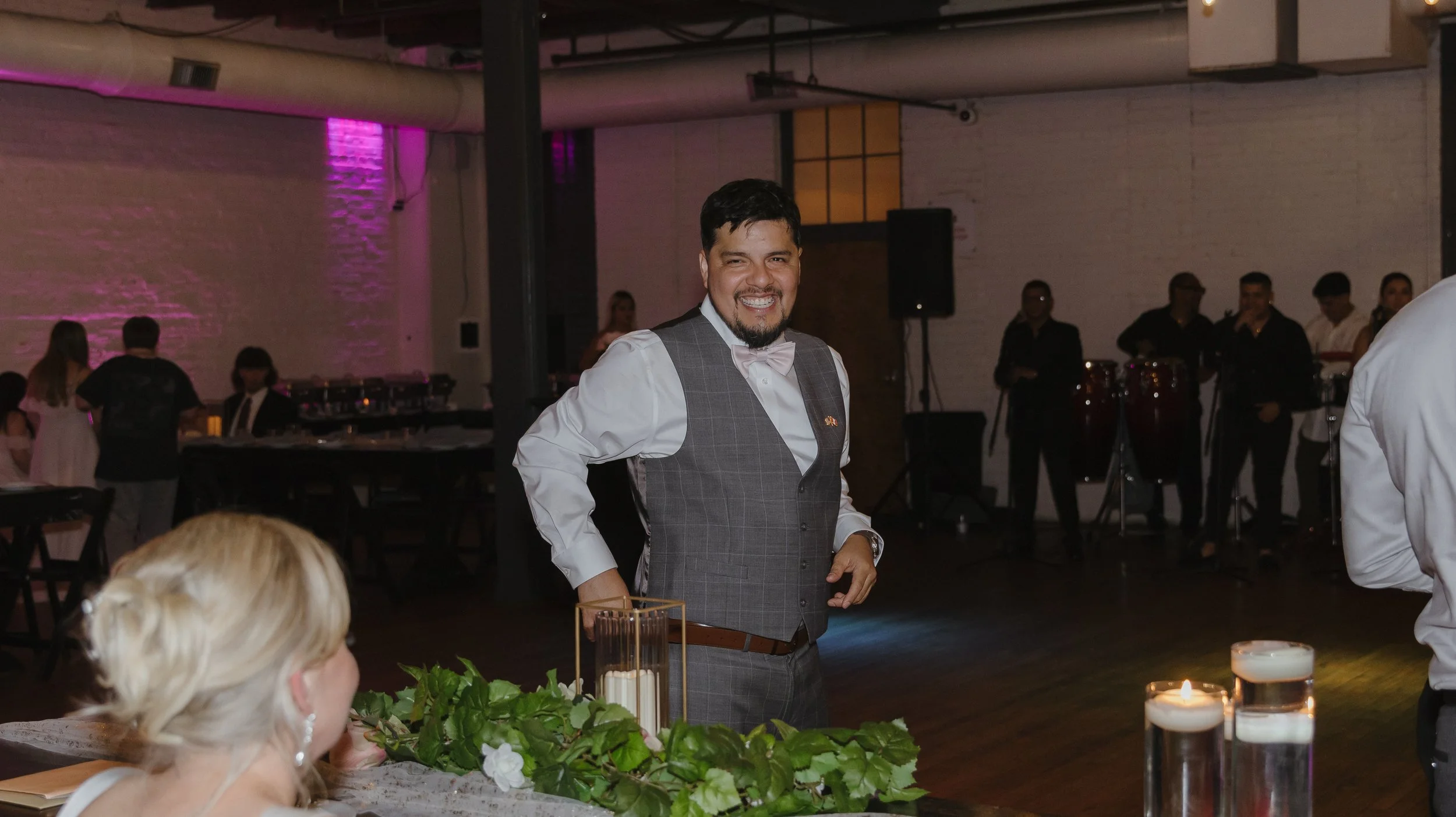 Smiling man in gray plaid vest with bowtie standing at a table decorated with greenery and candles at an indoor event.
