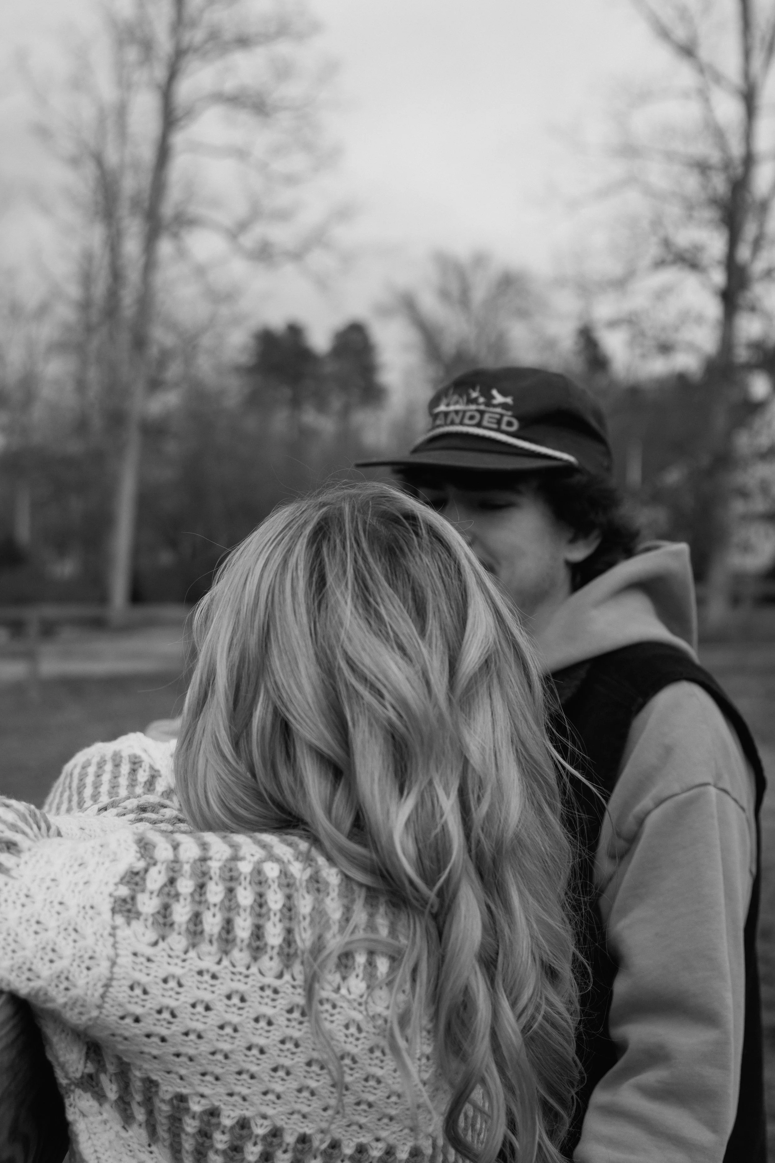 A black and white photo of two people embracing outdoors, their faces mostly obscured by the woman's hair and shoulder. The background shows leafless trees, indicating it might be fall or winter.