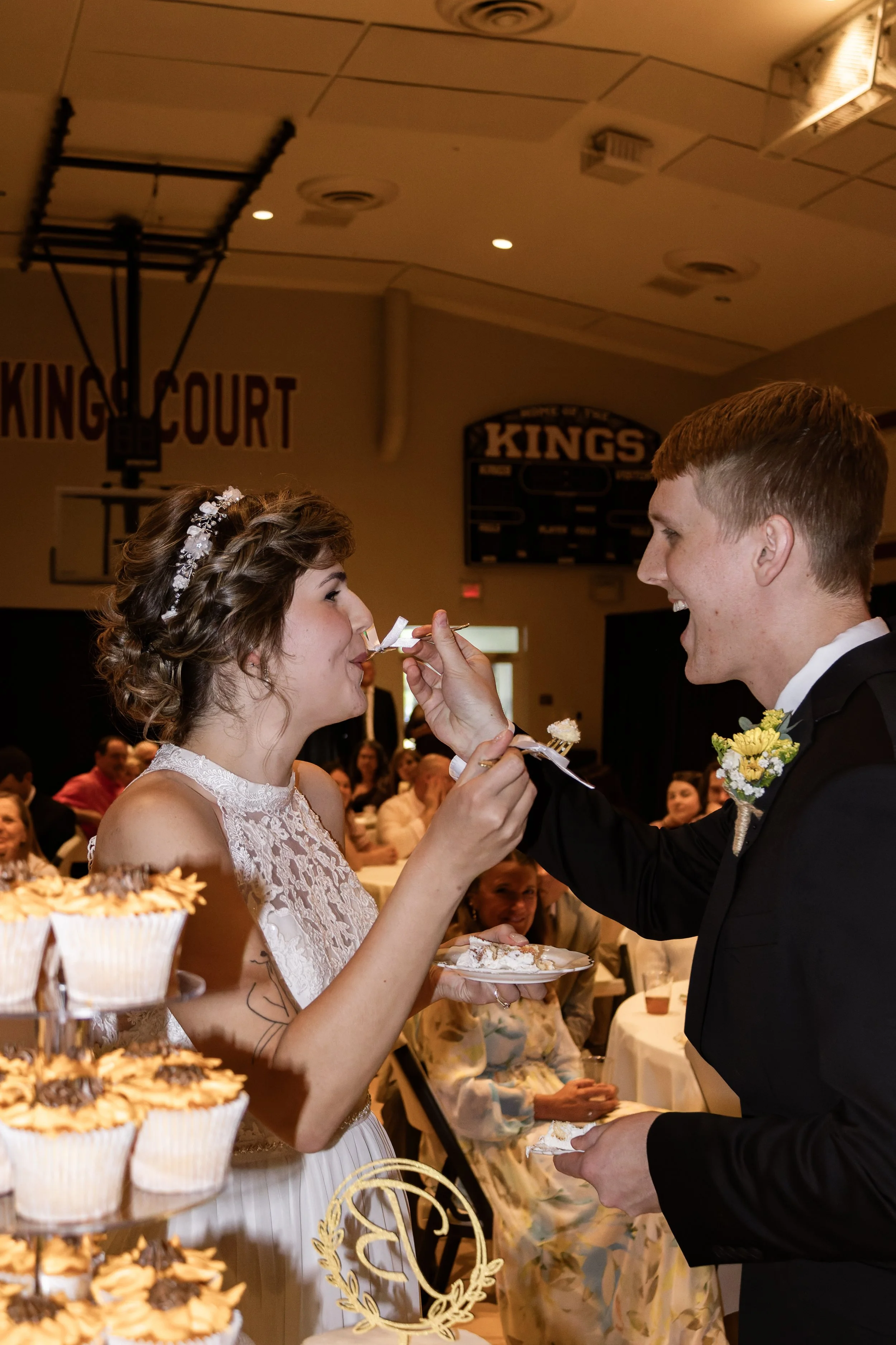 Couple at wedding reception sharing a piece of cake, with guests sitting at tables in the background.