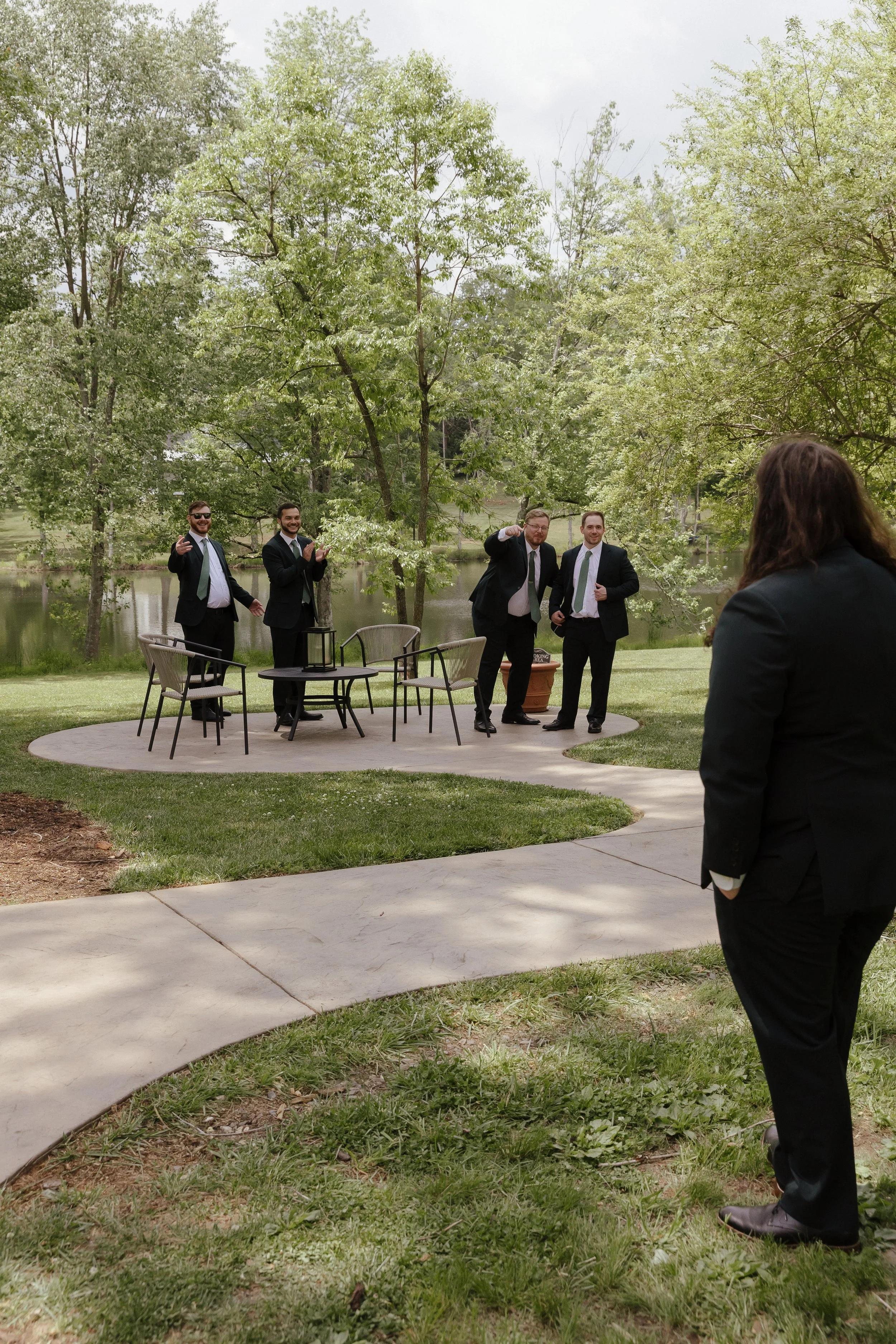 A photo of a group of 4 men in suits, celebrating when they see another man standing with  his back turned to the camera on a sunny day. 