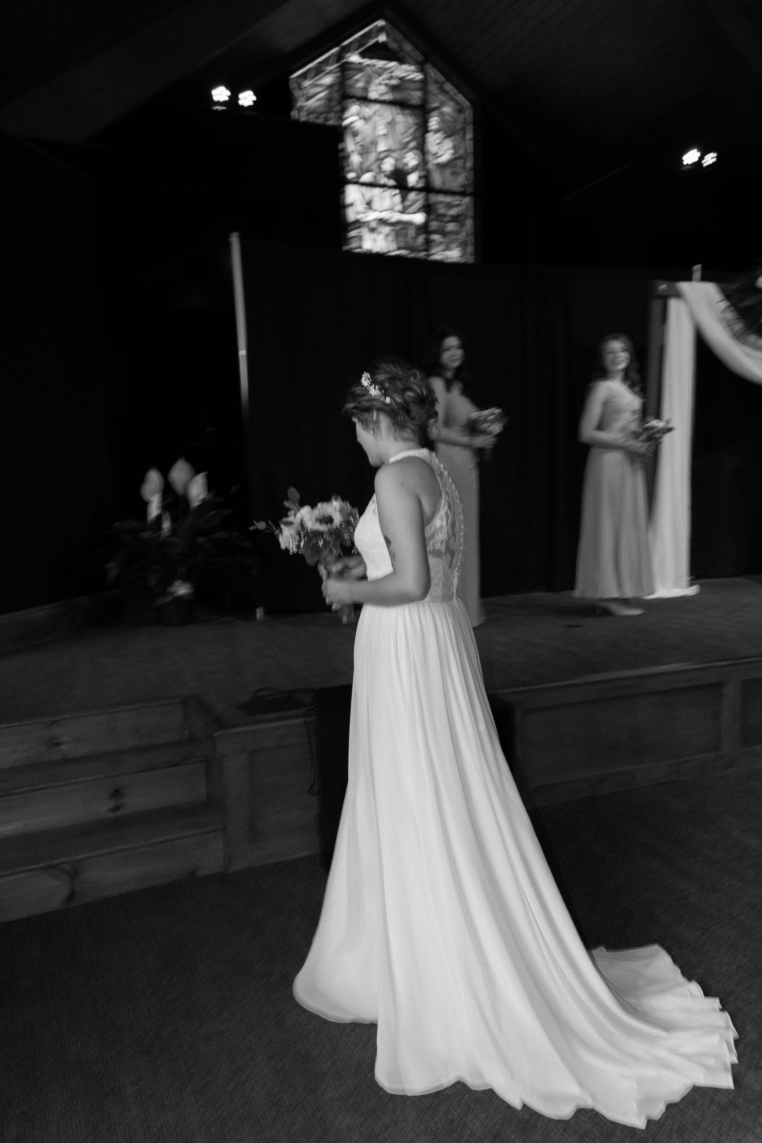 Black and white photo of a bride holding a bouquet, standing in front of three women on a stage, with a stained glass window in the background.