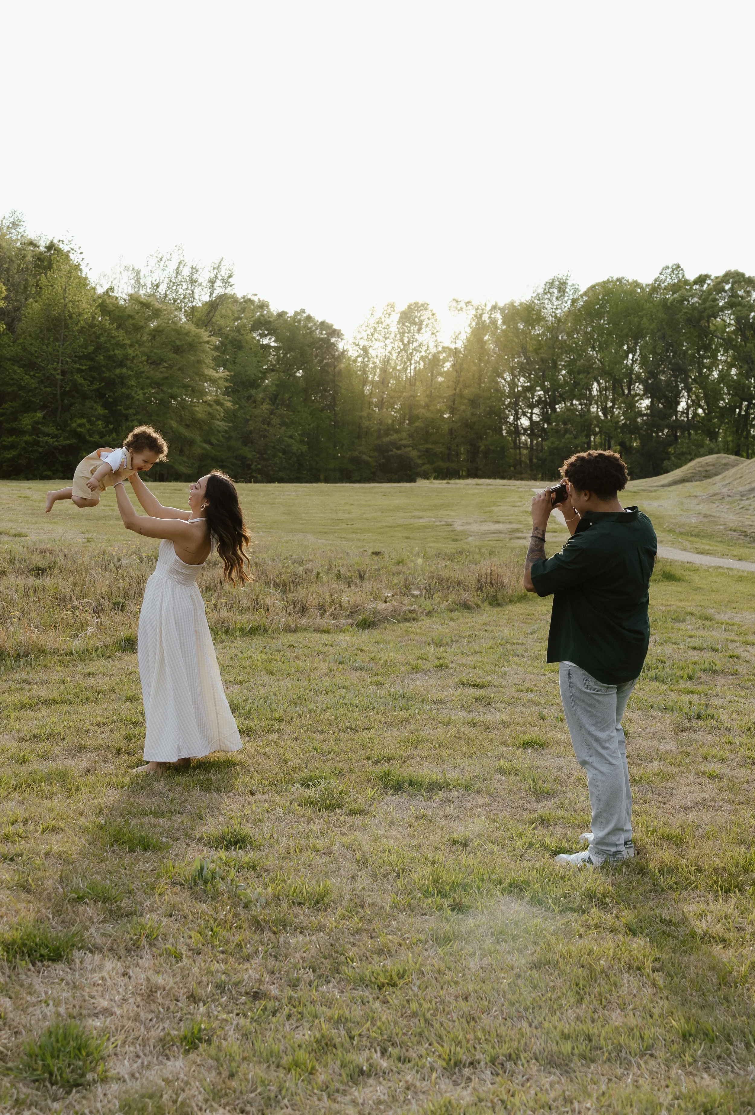 A woman in a white dress is playing with a young child in a field while a man takes a photo of them with a camera. The scene is set outdoors during sunset with trees in the background.