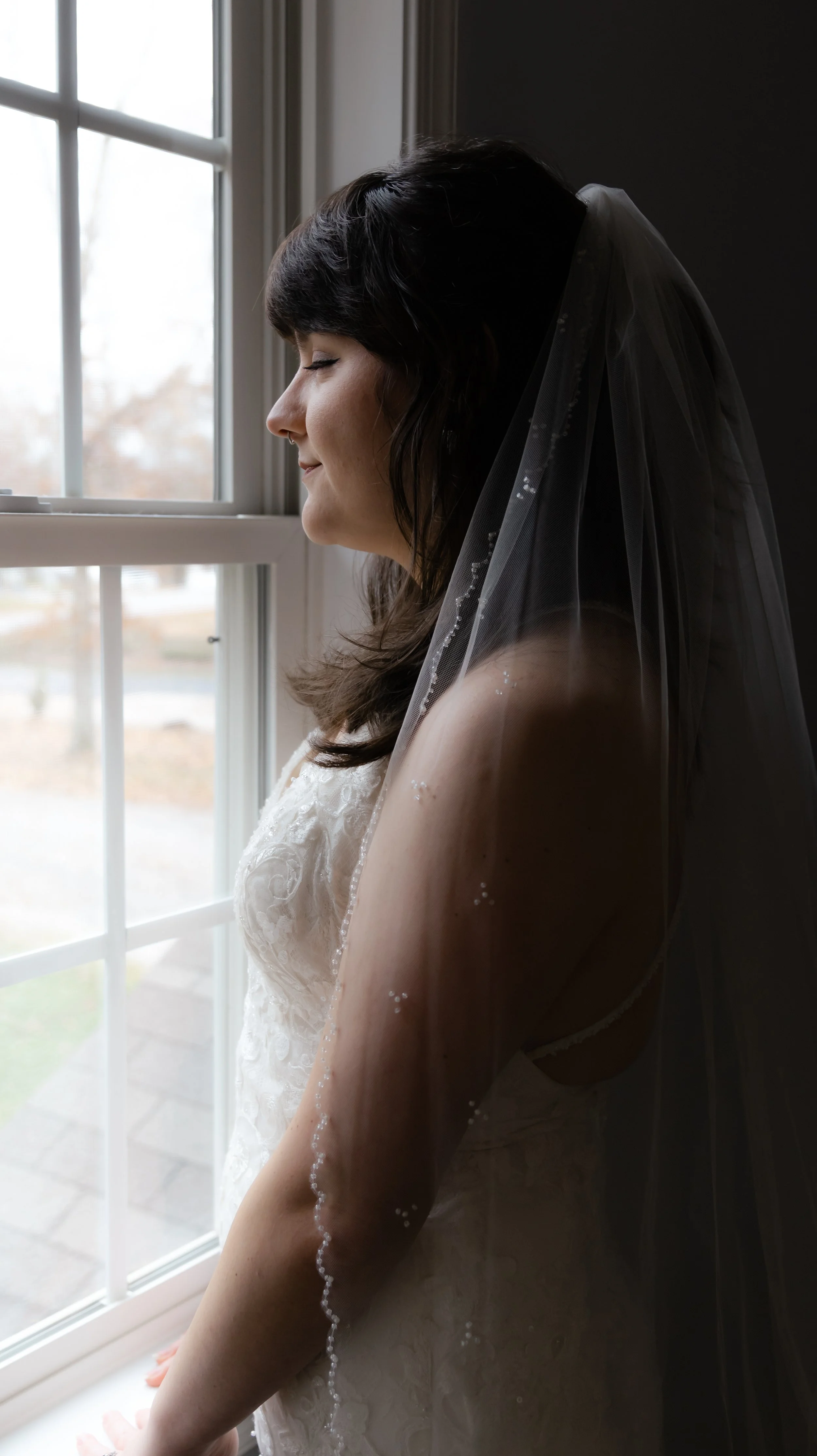 Bridal woman with dark hair and veil, standing by a window with closed eyes and a peaceful expression.