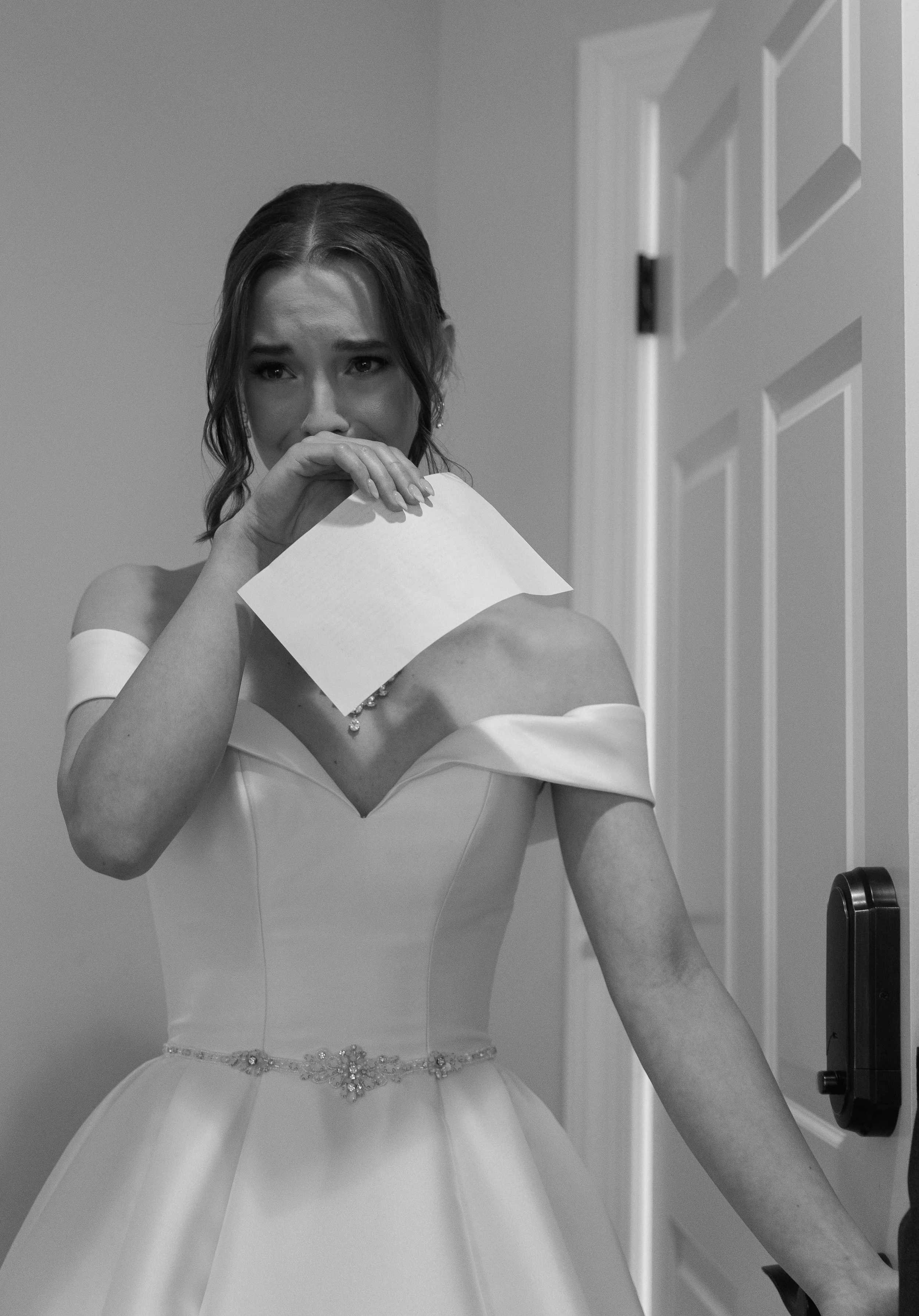 A woman in a wedding dress, holding a piece of paper, standing in front of a mirror in a fitting room, wiping away tears.