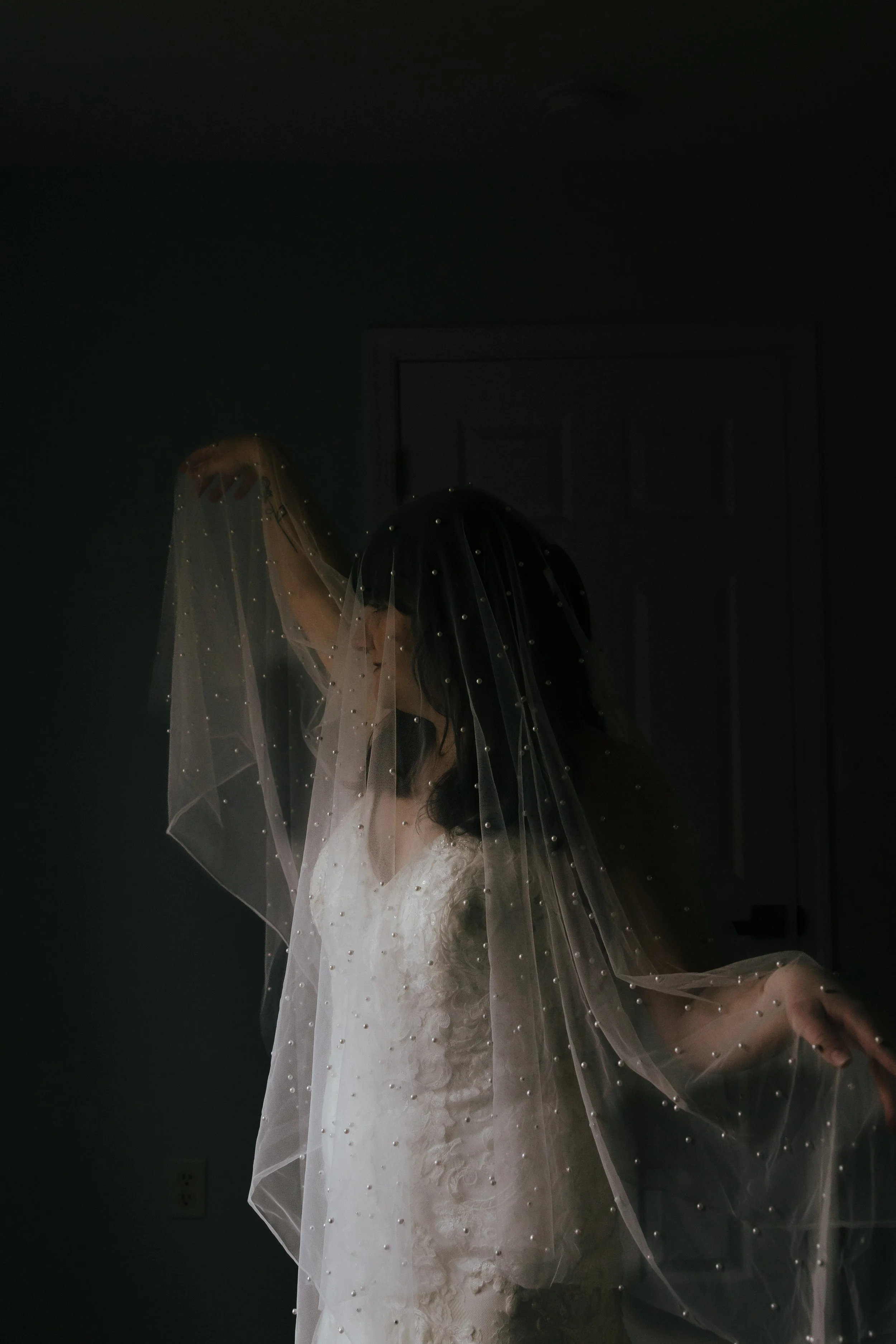 A bride wearing a white wedding dress and a veil with pearl accents, standing in a dimly lit room, with her face partially obscured by the veil.
