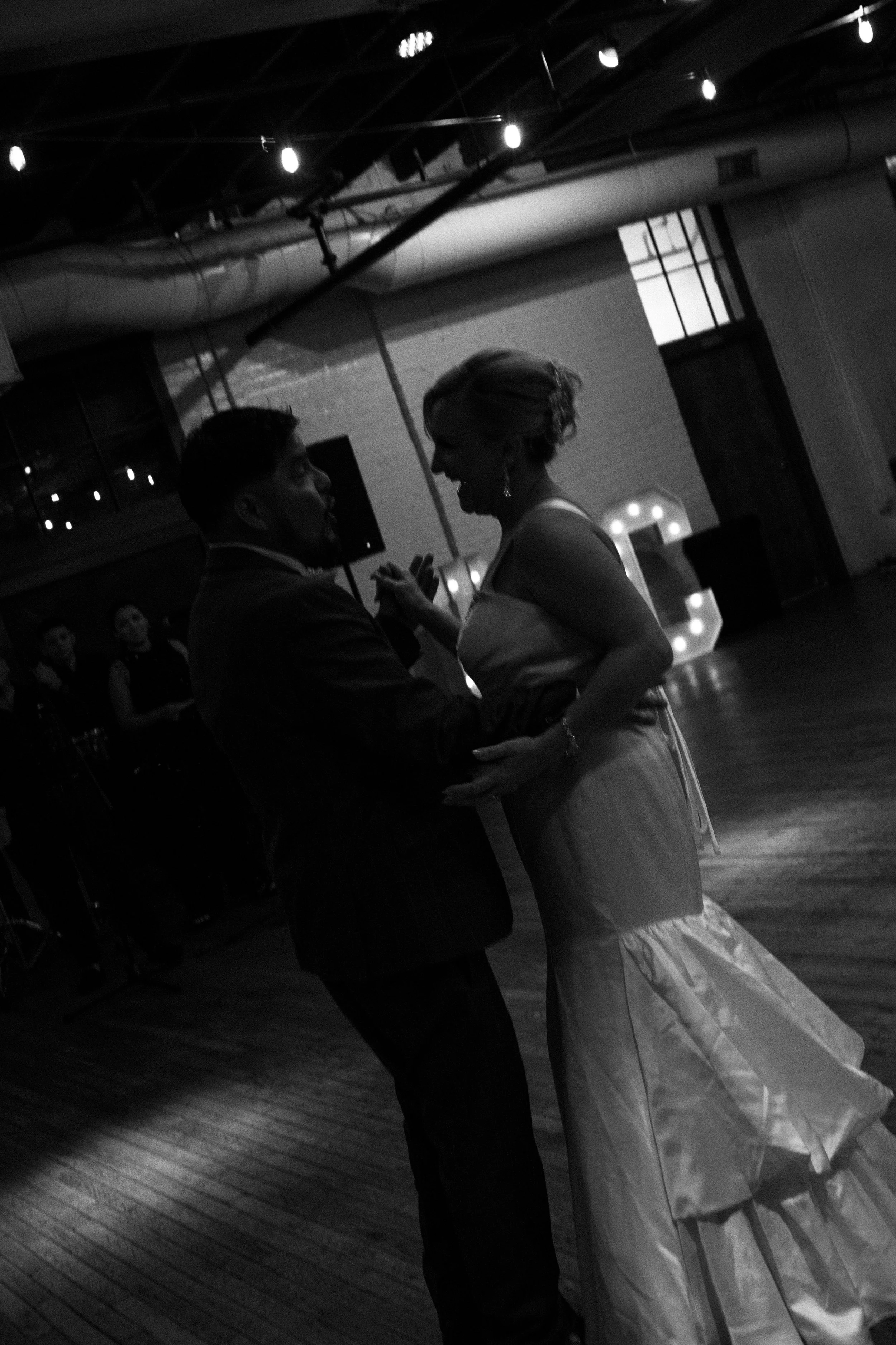Black and white photo of a couple dancing closely at a wedding reception, with guests and a large illuminated letter in the background.
