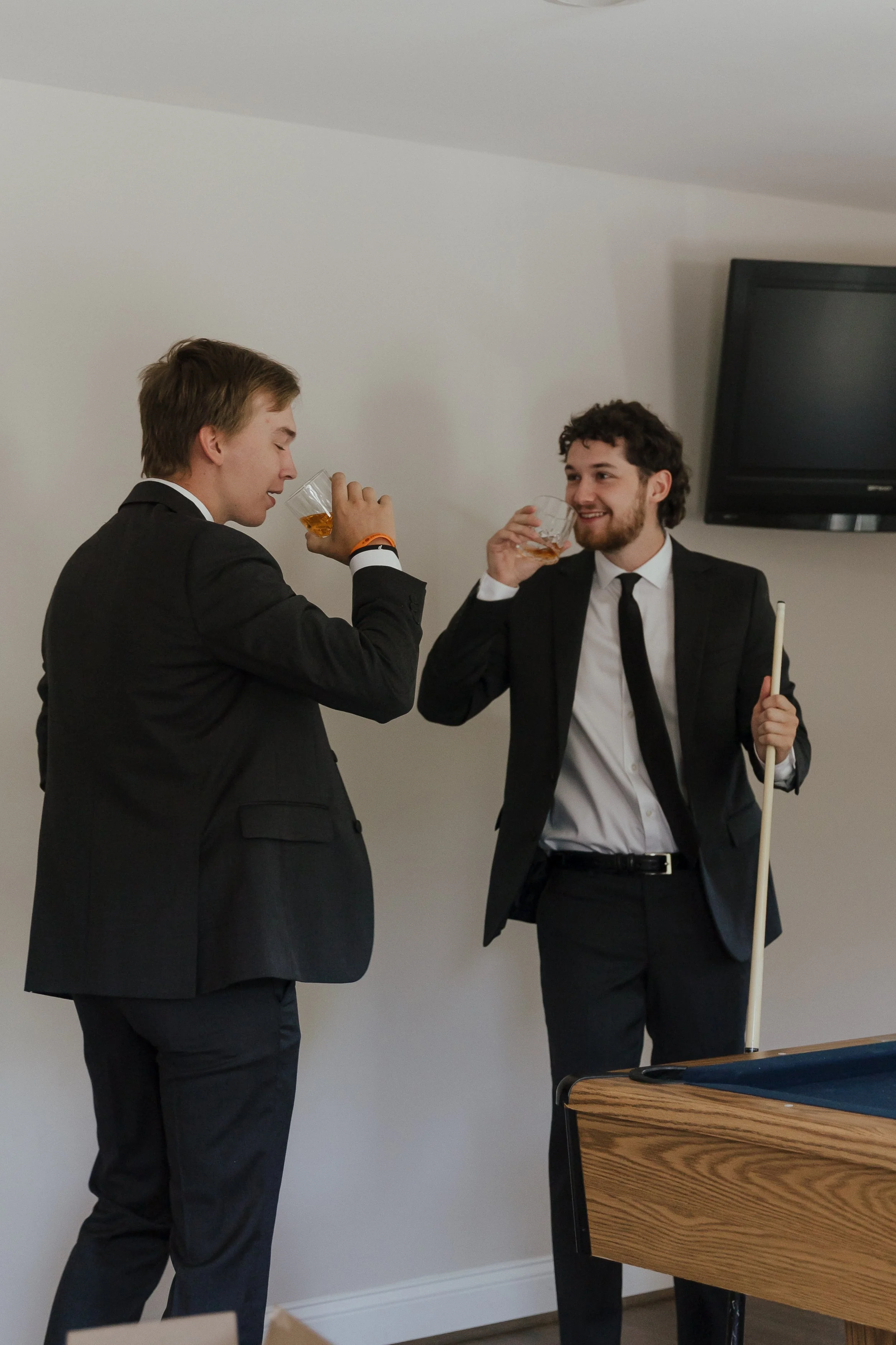 Two young men in suits enjoying drinks and conversing in a room with a pool table and a TV mounted on the wall.